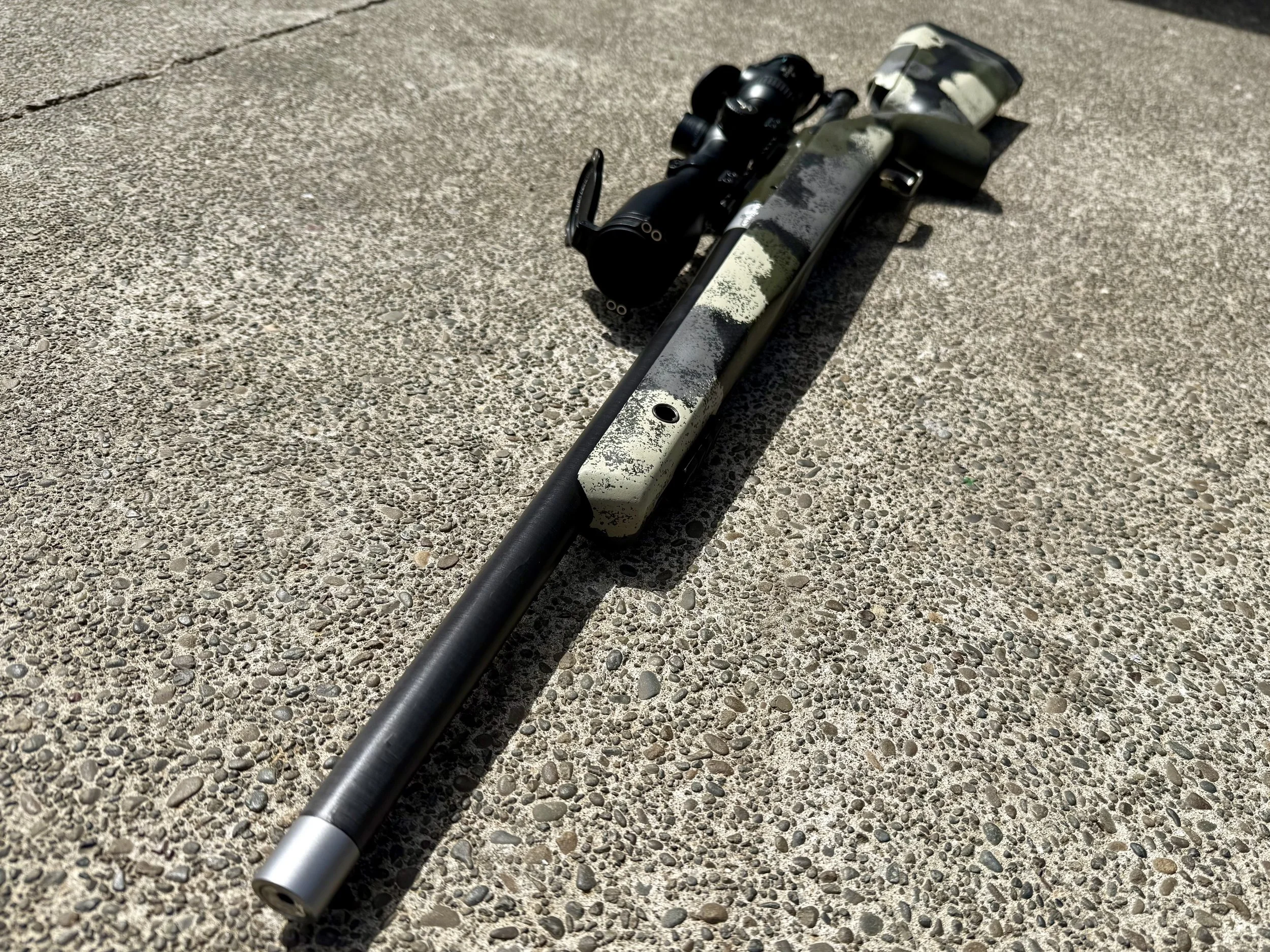 A sniper rifle with a camouflage pattern lying on a pebbled concrete surface.