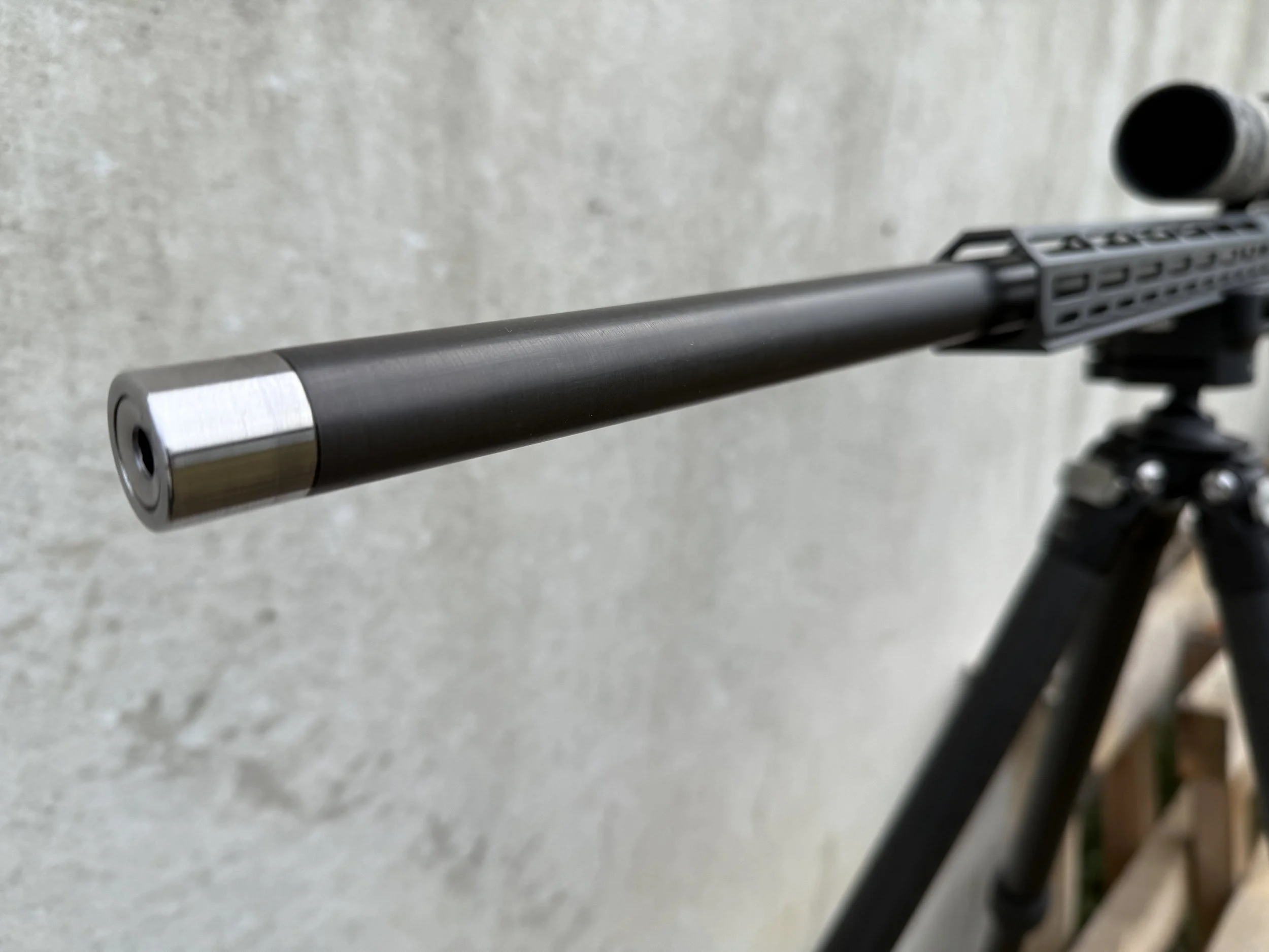 Close-up view of a firearm barrel and part of a mounted scope or rail system set against a light gray concrete background.