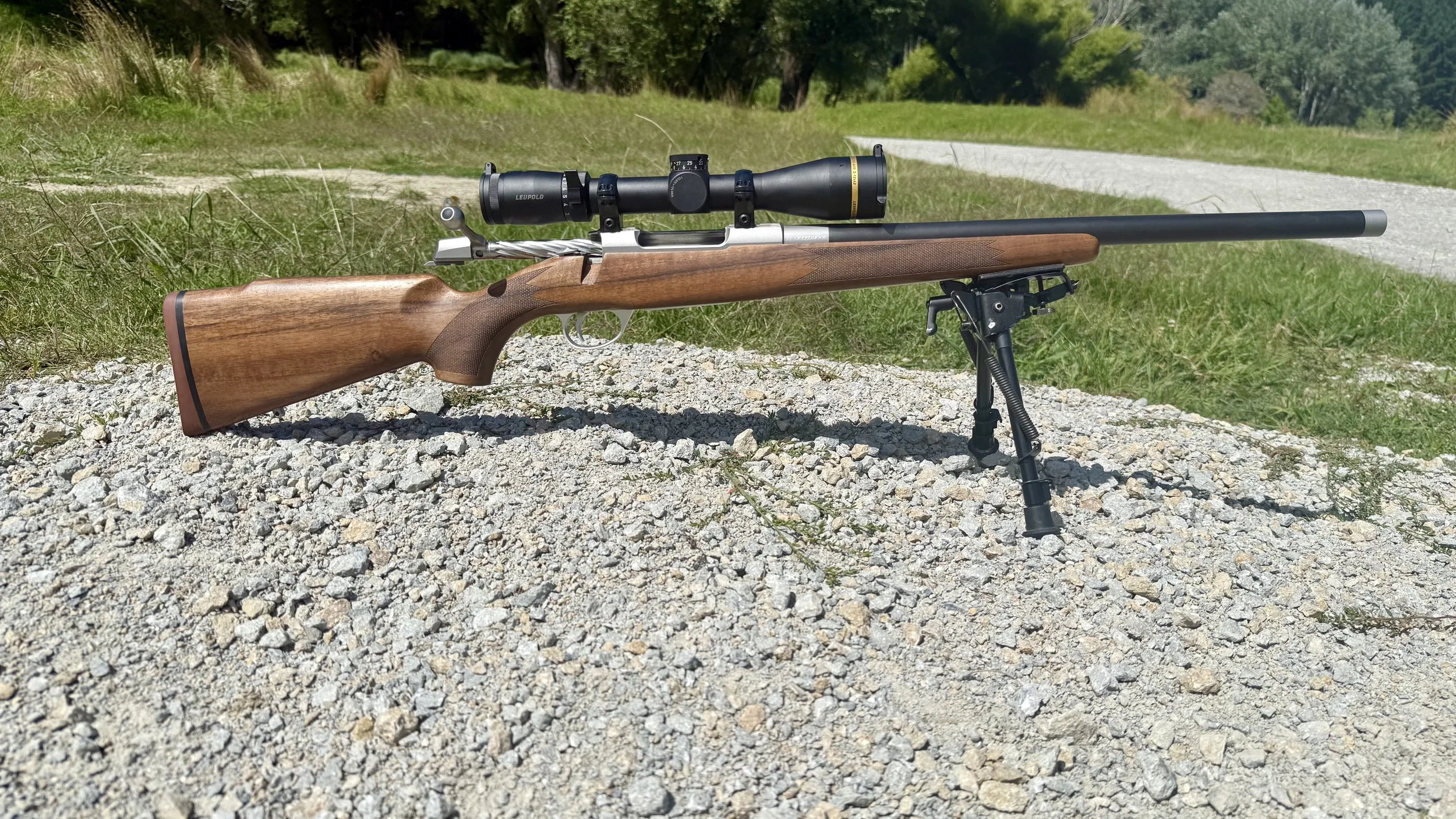 A rifle with a wooden stock, mounted with a scope, rests on small rocks outdoors on a sunny day with grass, trees, and a gravel path in the background.