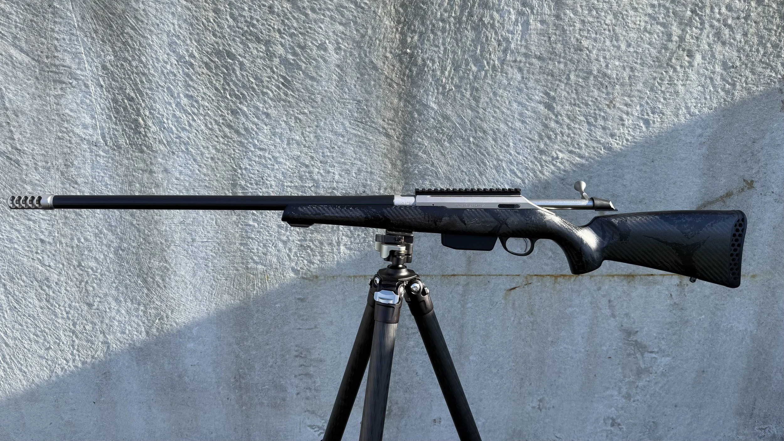 A rifle with a scope mounted on a tripod, resting on a textured gray concrete surface.