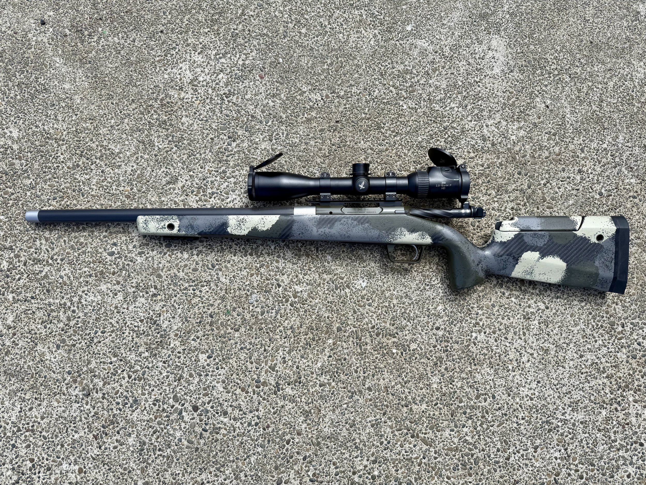 A scoped rifle with a camouflage pattern on the stock rests on a gravel surface.
