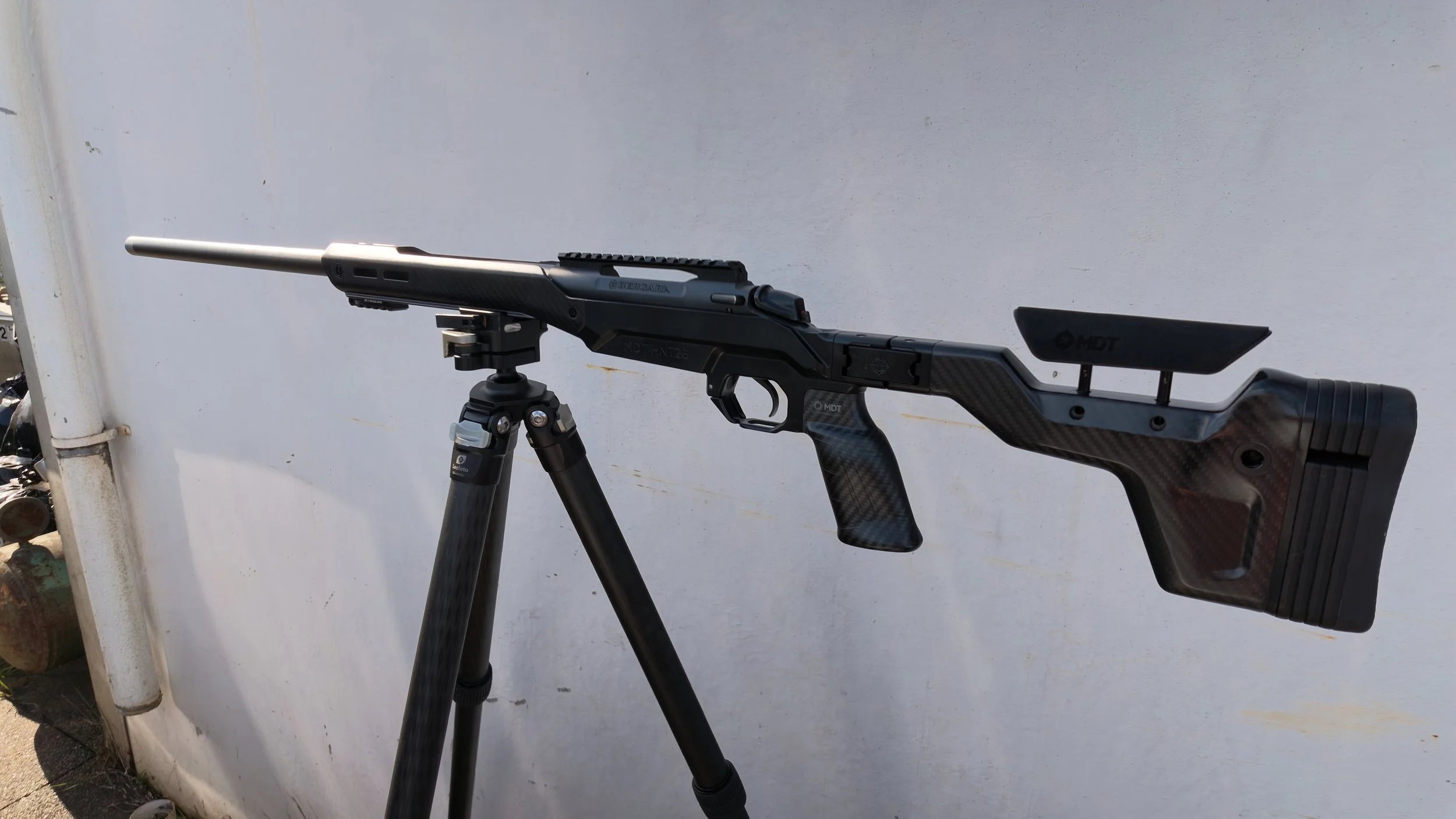 A black racing gun mounted on a tripod stand against a plain white wall.