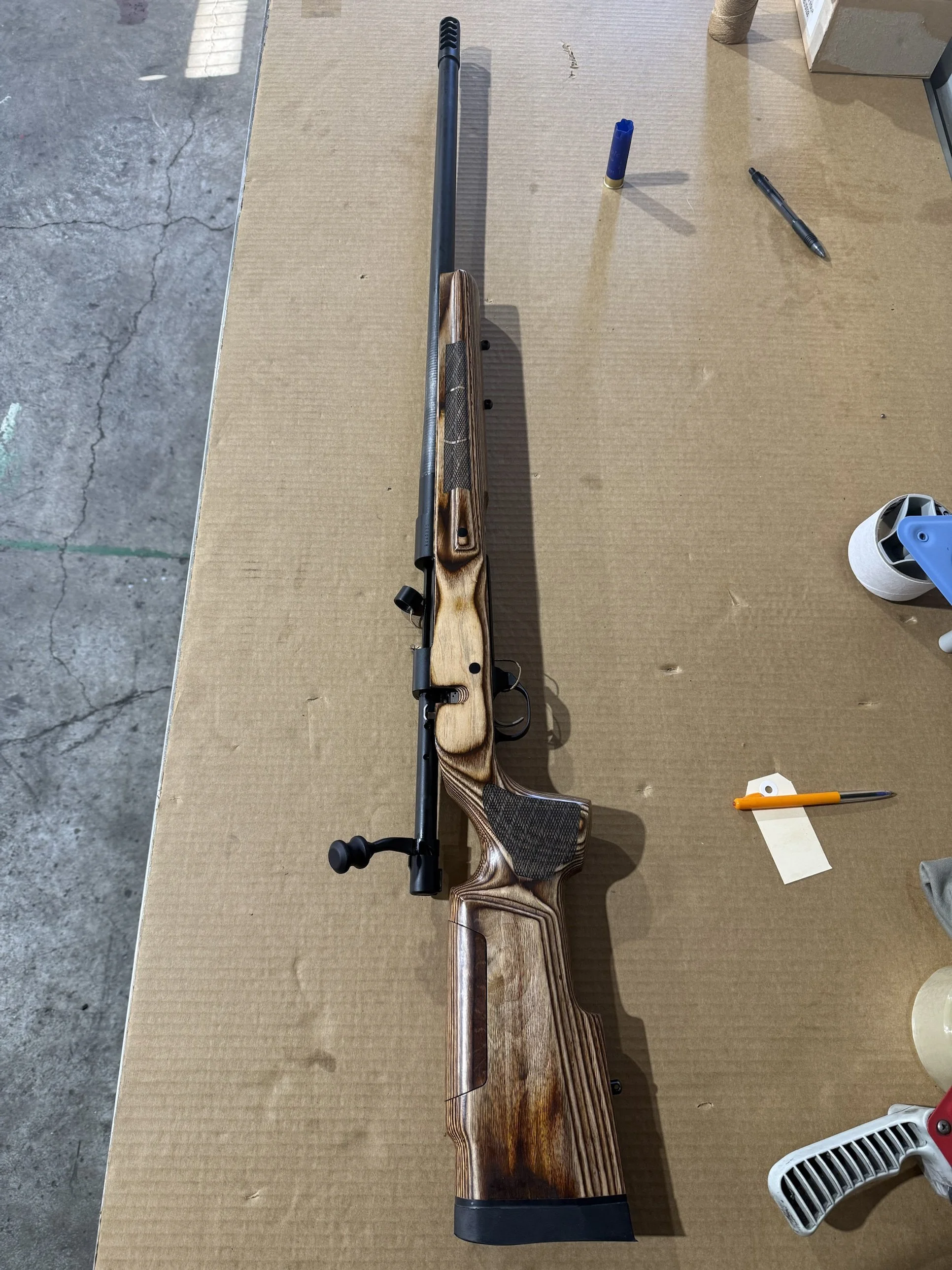 A bolt-action rifle with a wooden stock and black barrel resting on a brown textured surface, with a marker, a blue container, and a roll of tape nearby.