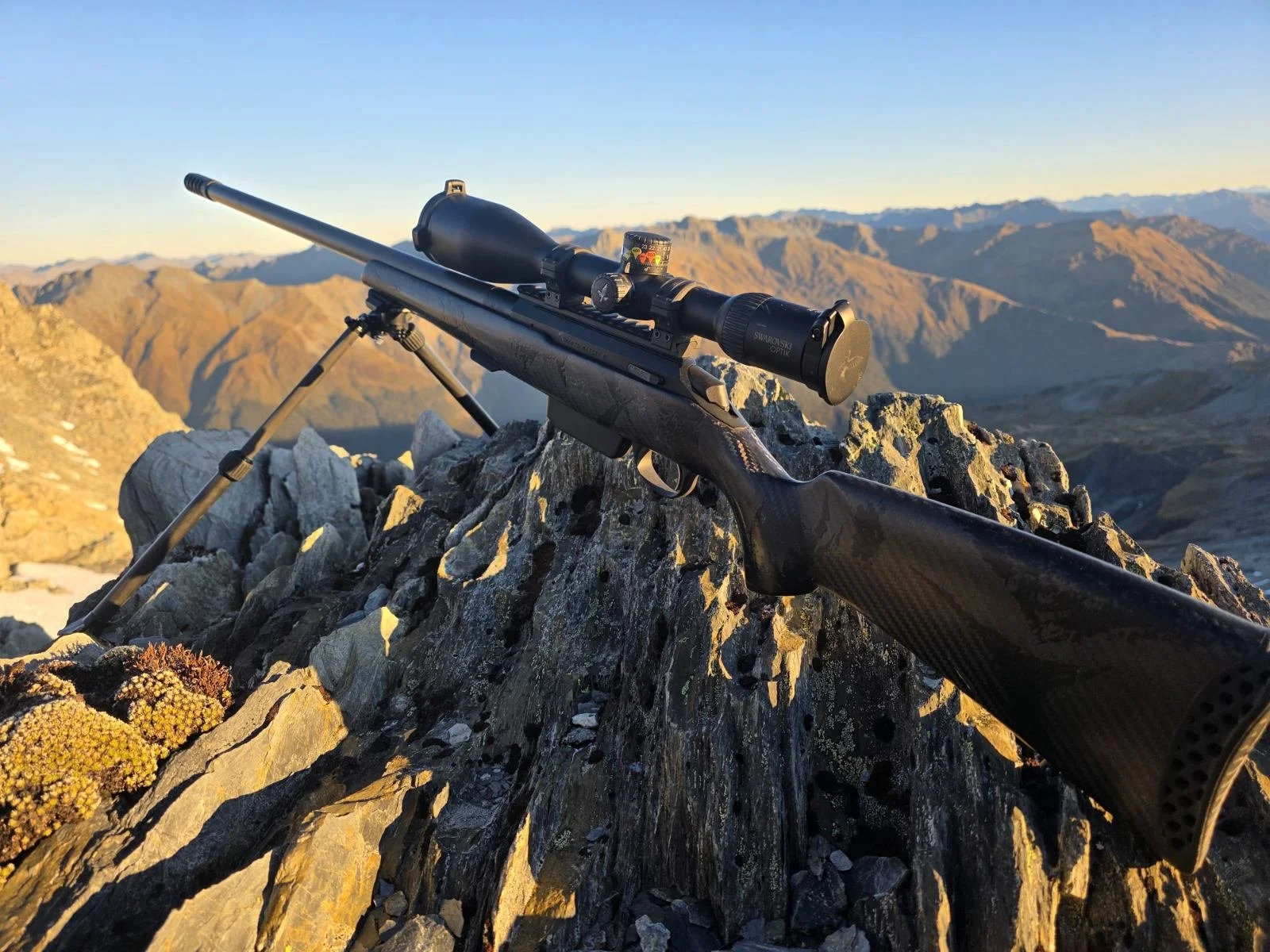 A sniper rifle with a scope is resting on a rocky mountain summit, overlooking a vast mountain range with rugged peaks in the background during sunset.
