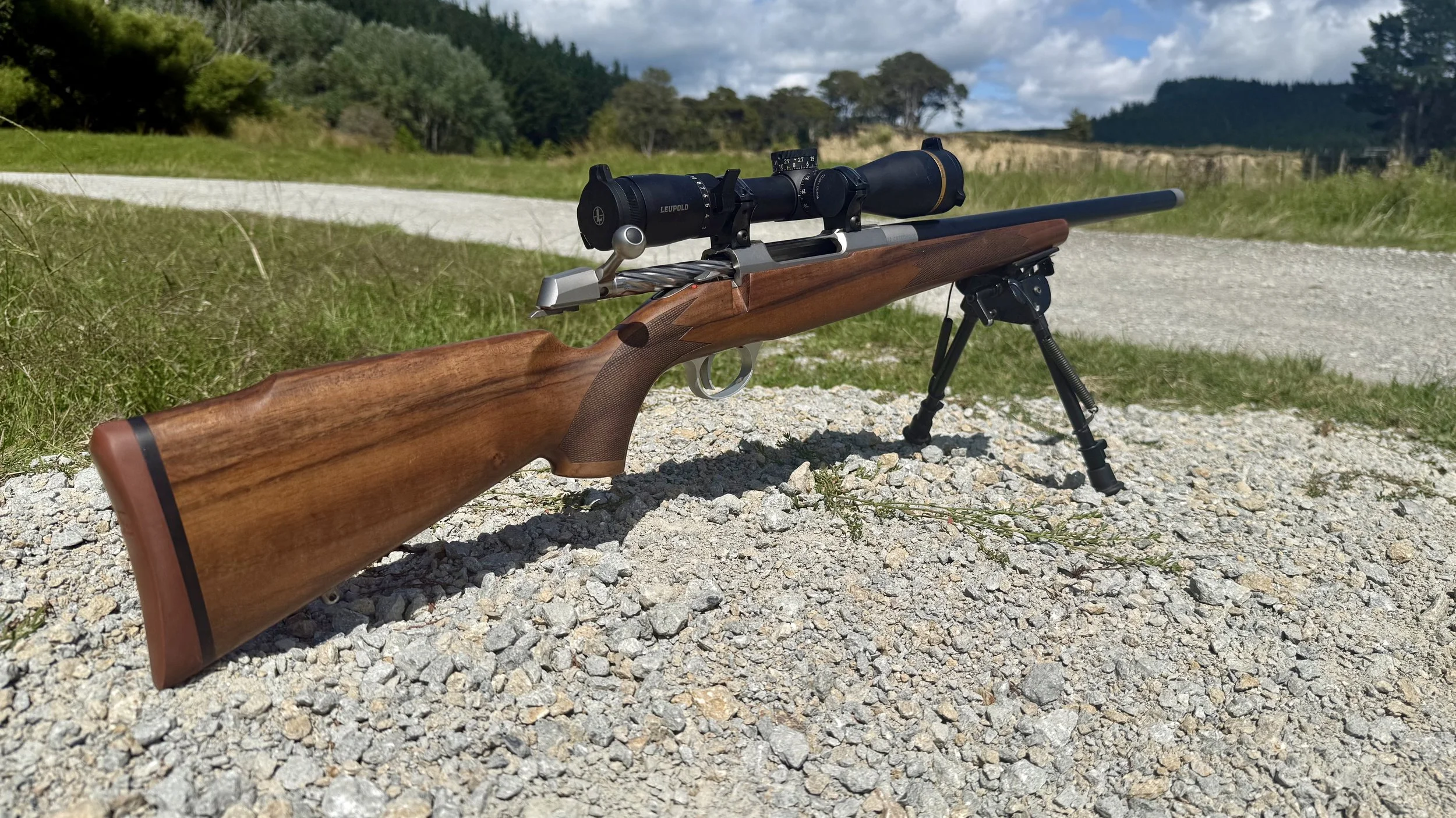 A bolt-action rifle with a wooden stock and a mounted scope, set on a bipod outdoors on rocky ground with grass and trees in the background.