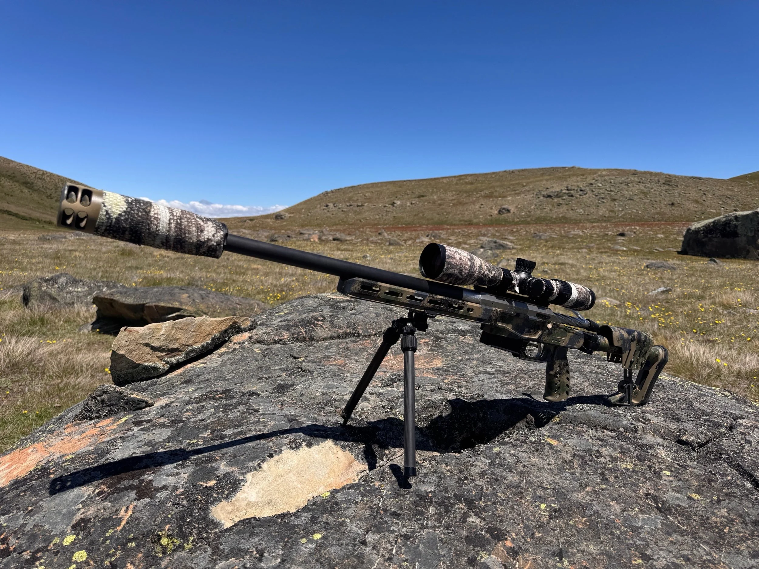 A varmint scope lying on a rocky outdoor terrain with hills and a clear blue sky in the background.