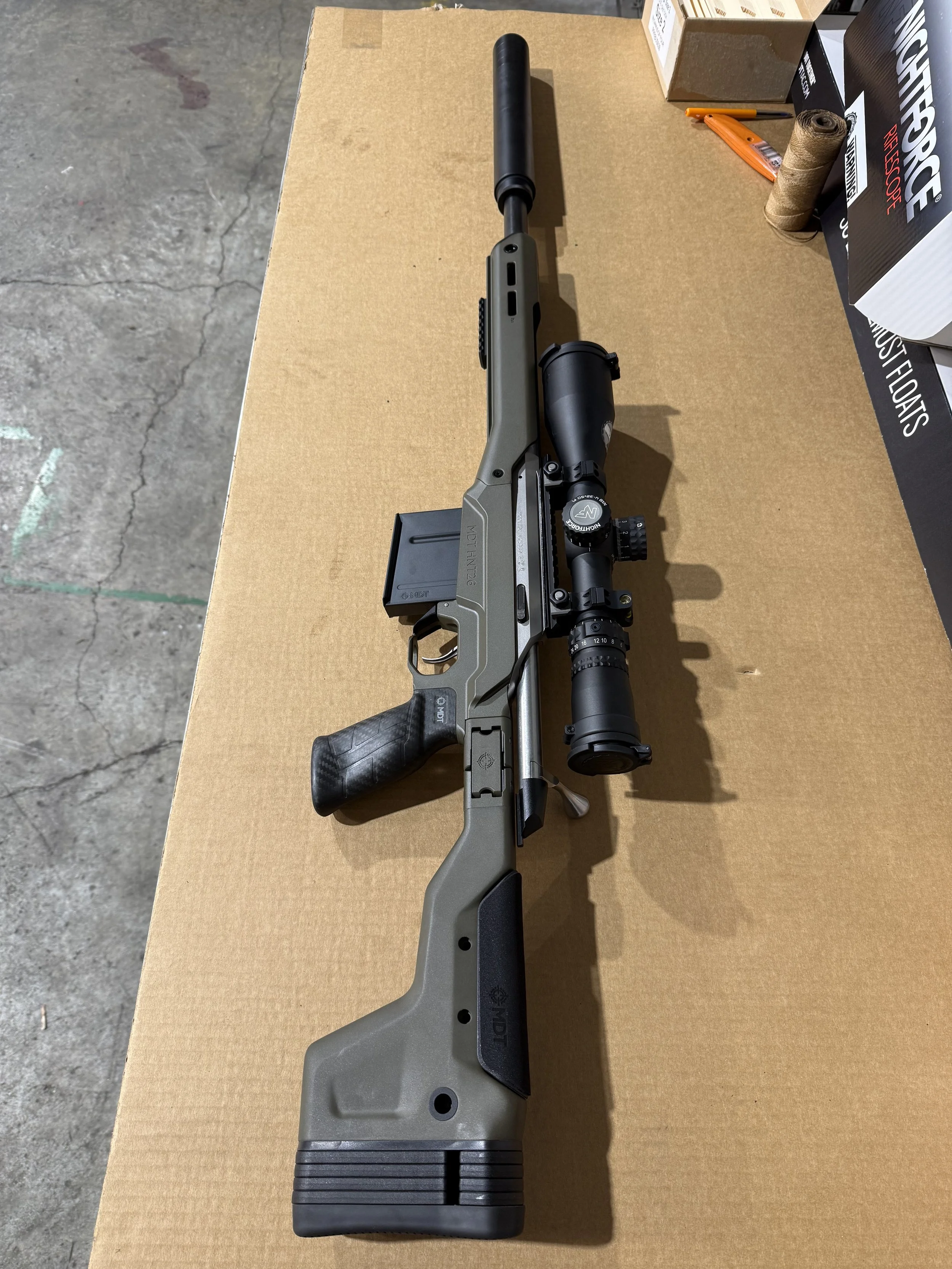 A rifle with a scope and a black stock is placed on a cardboard surface, with various boxes and tools nearby.