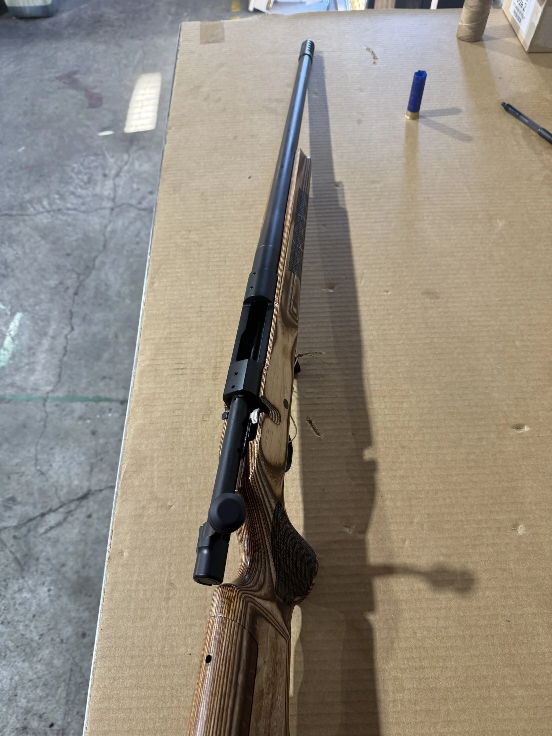A shot of a rifle with a wooden stock lying on a piece of cardboard in an indoor setting, with some markers or pens nearby.