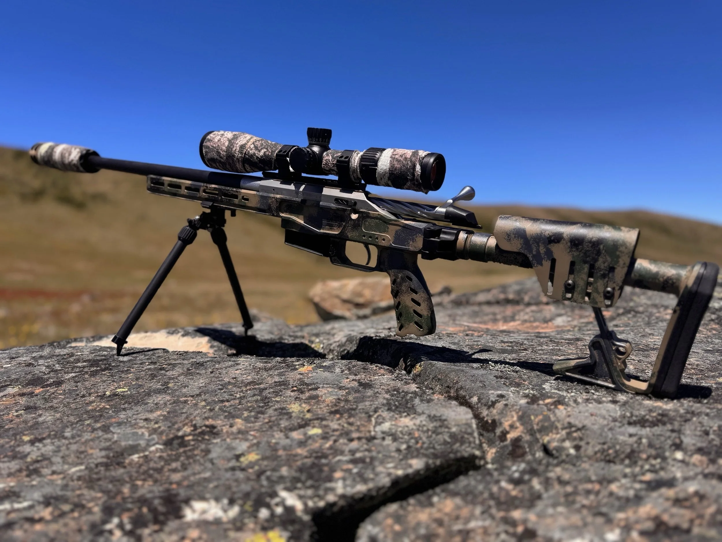 A scoped rifle with camouflage pattern stored on a bipod on a rock outdoors against a clear blue sky.