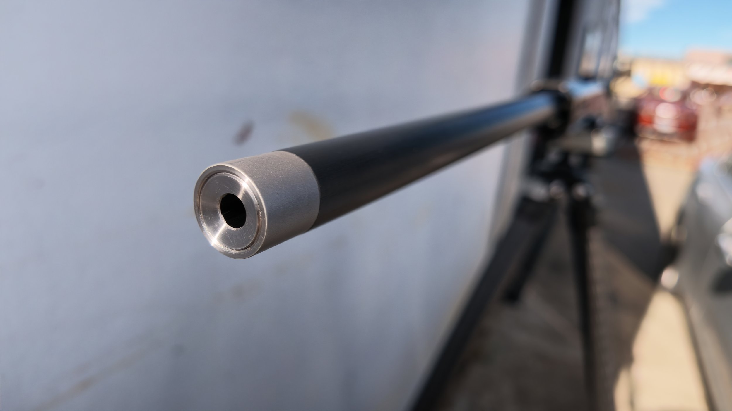 Close-up of a metallic gun barrel mounted on a tripod, with a blurred urban background.