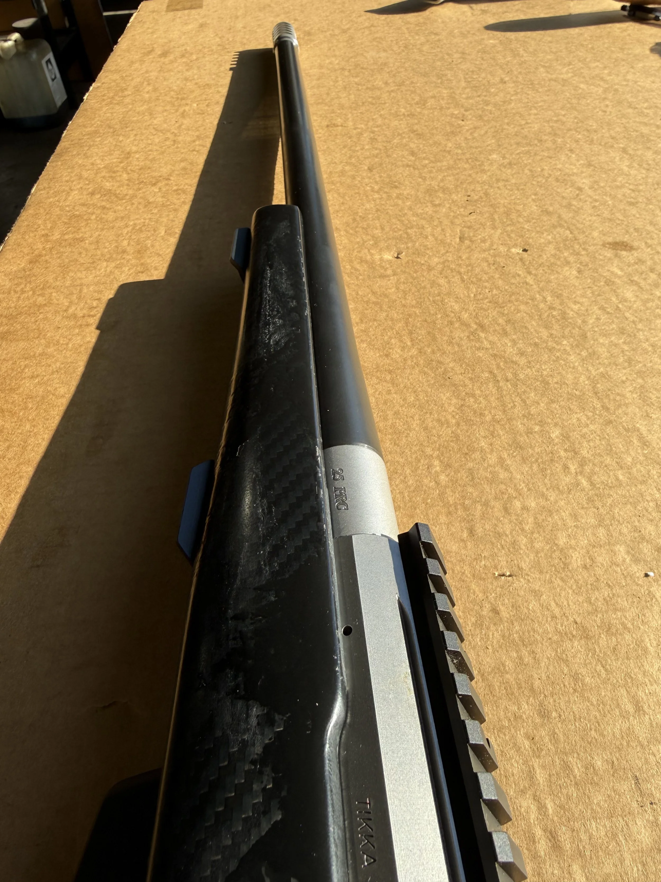 A black and silver rifle with a long black barrel placed on a brown surface, seen from a top-down perspective. The focus is on the barrel and magazine of the firearm.