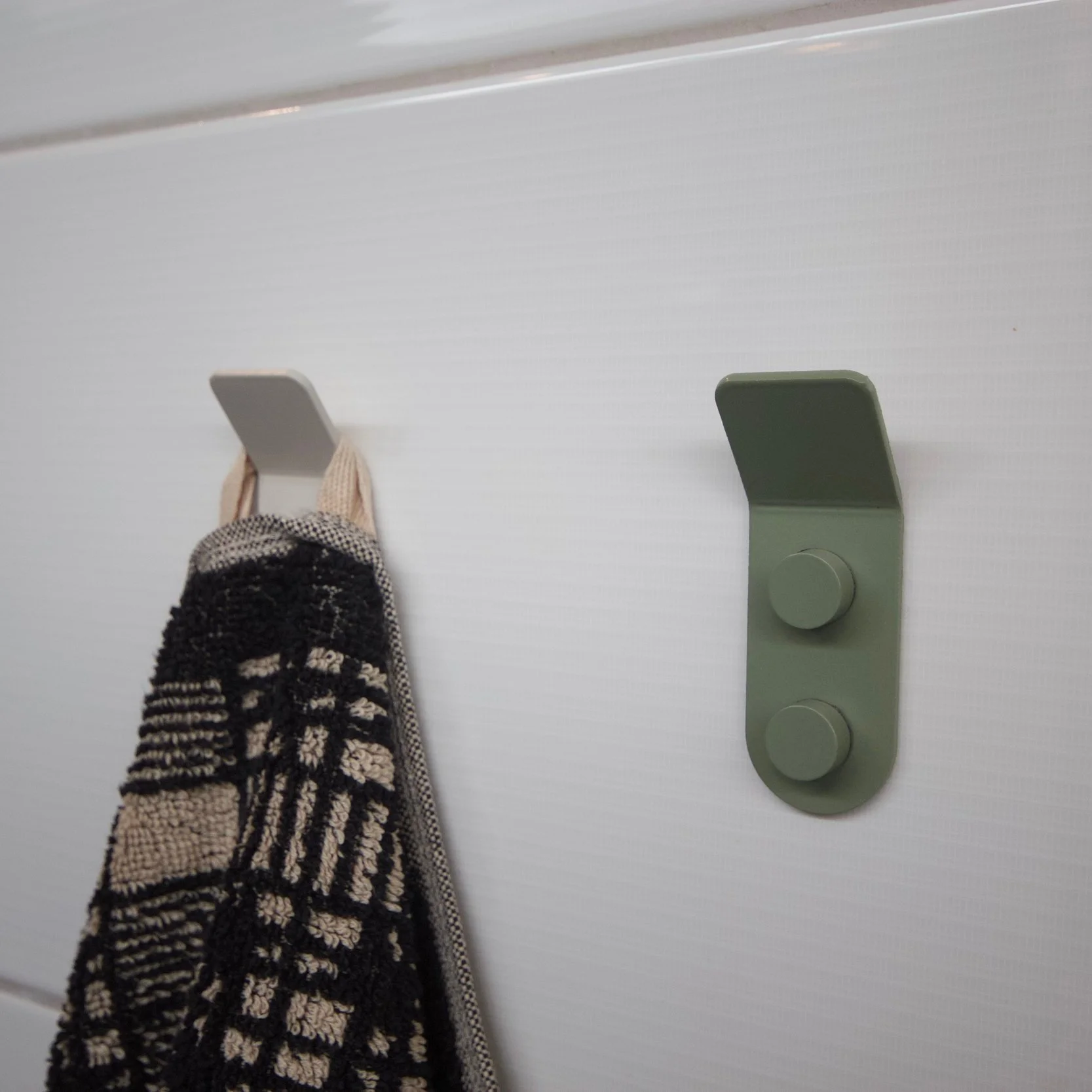 A black and beige checkered towel hanging on a white wall hook next to a green wall hook with two knobs.