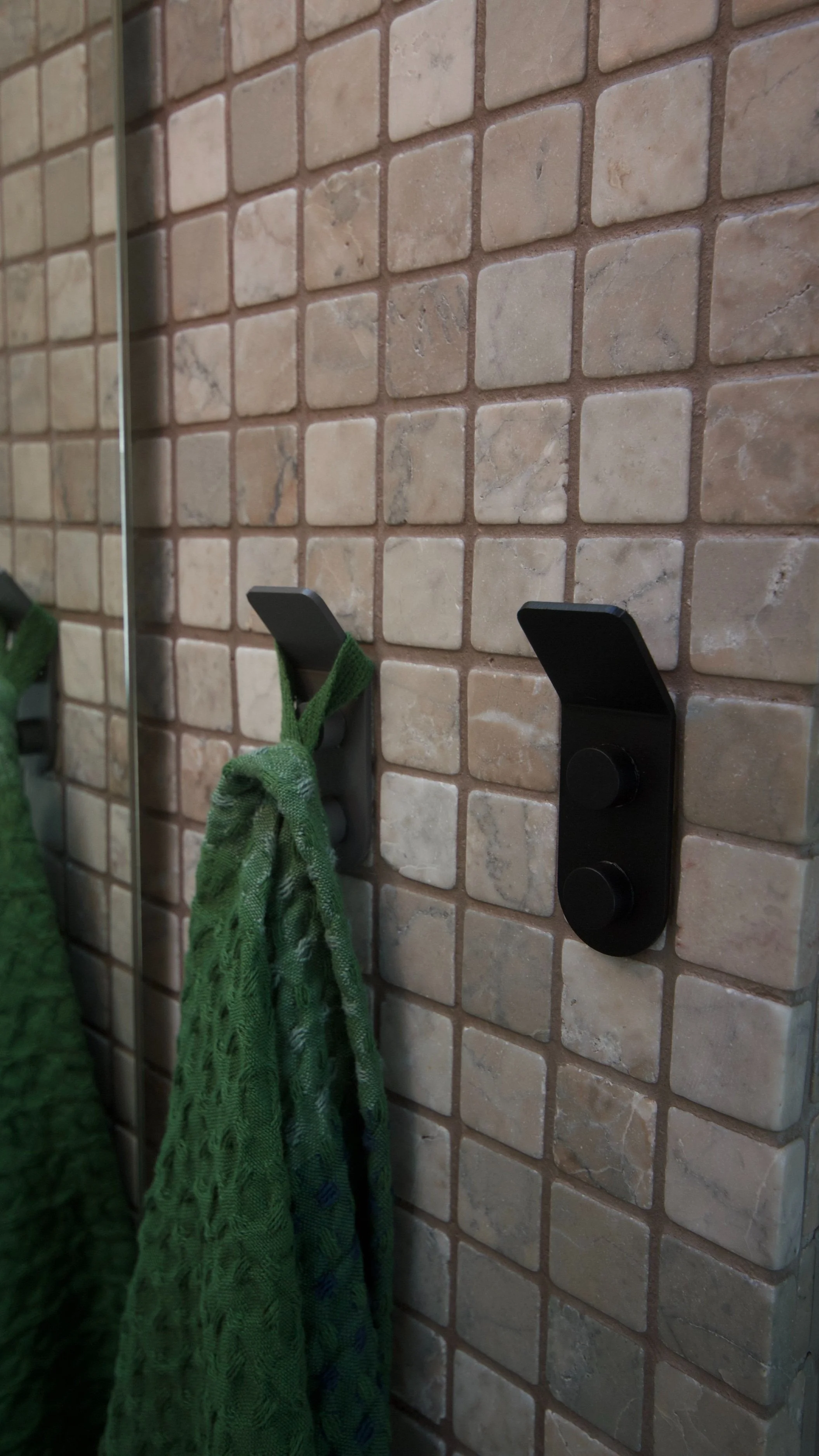 Close-up of a wall with beige square tiles, showing two black wall hooks, one with a green cloth hanging.