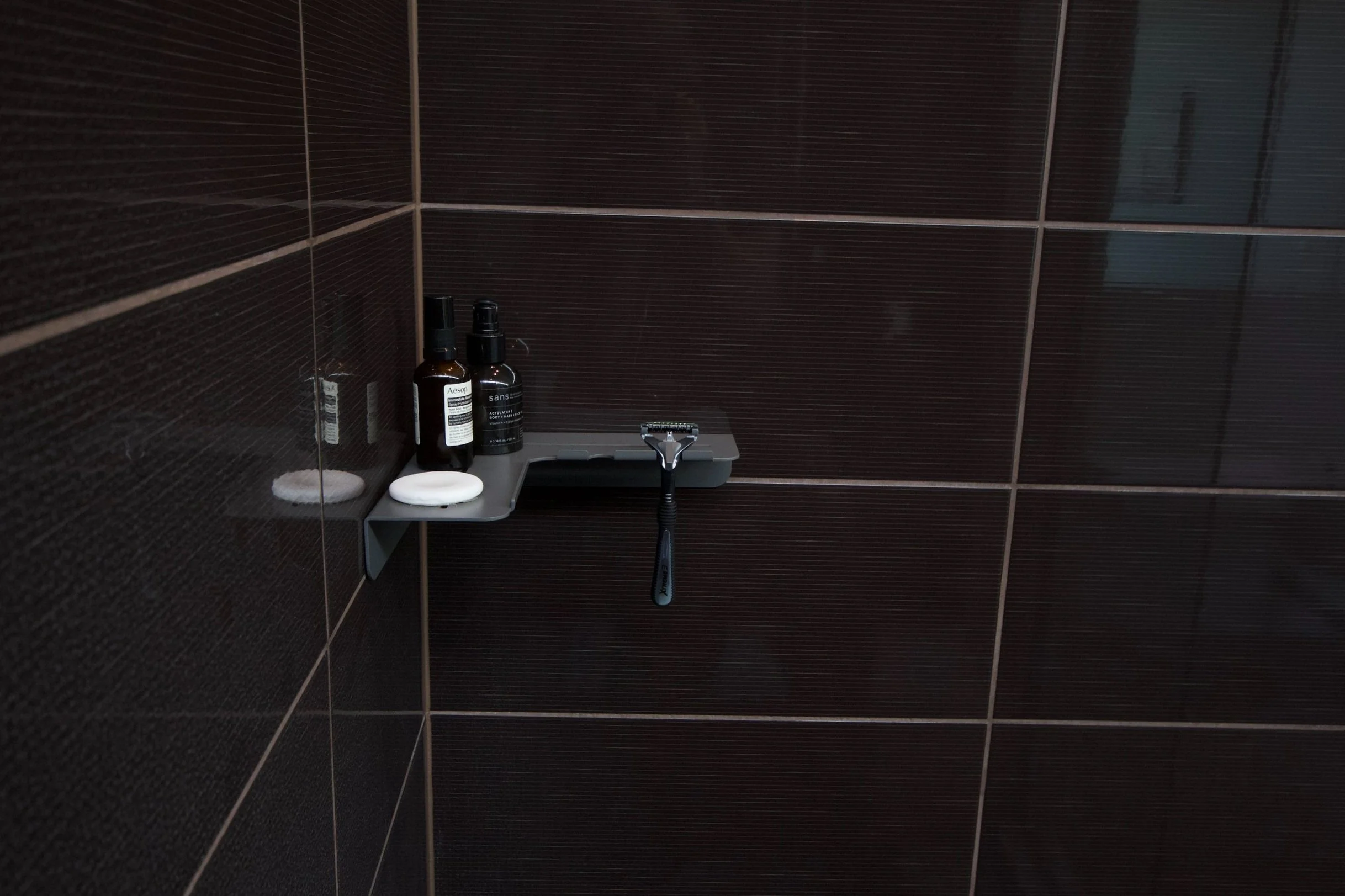 Close-up of a bathtub corner with a showerhead, dark brown tiled wall, and a small shelf holding bottles of toiletries, soap, and a razor.