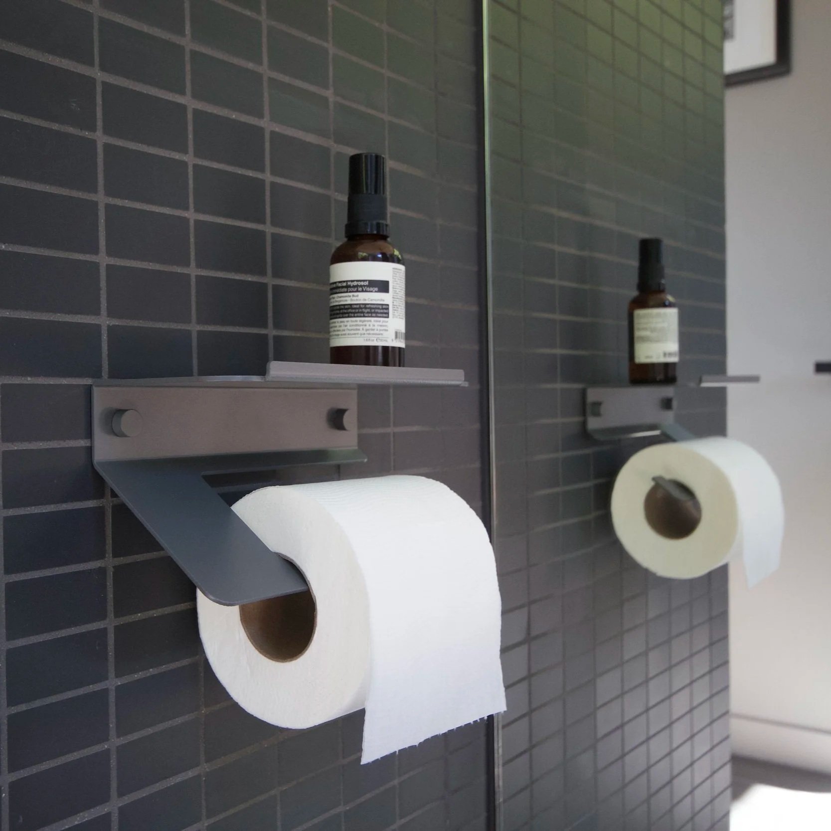 Minimalist grey aluminum toilet roll holder with an integrated shelf, mounted on a dark grey tiled wall, holding a toilet roll and a small bottle on the shelf, reflected in a mirror.