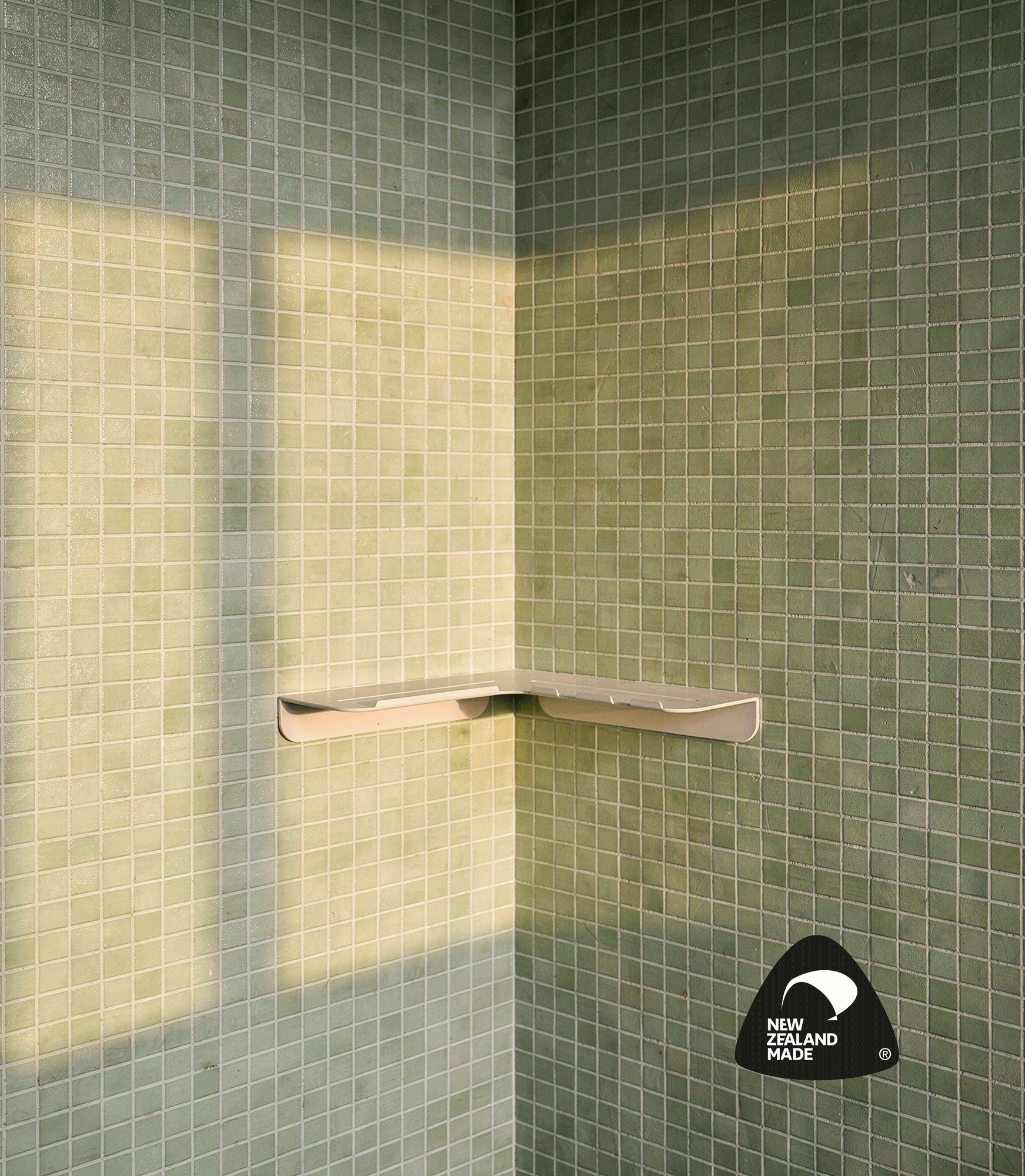A corner of a tiled shower with green square tiles, a white soap dish mounted on the wall, and a black sticker with 'New Zealand Made' in white text at the bottom right corner.