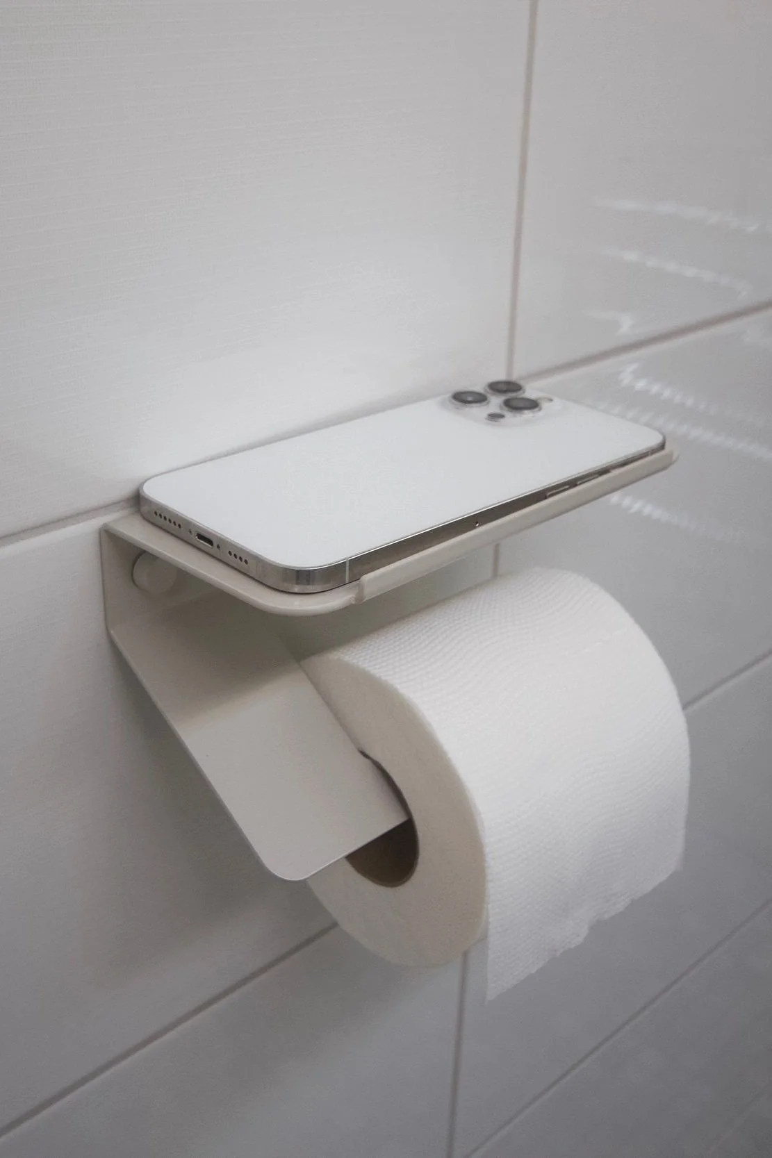 A smartphone placed on a white shelf above a toilet paper roll in a bathroom with white tiled wall.