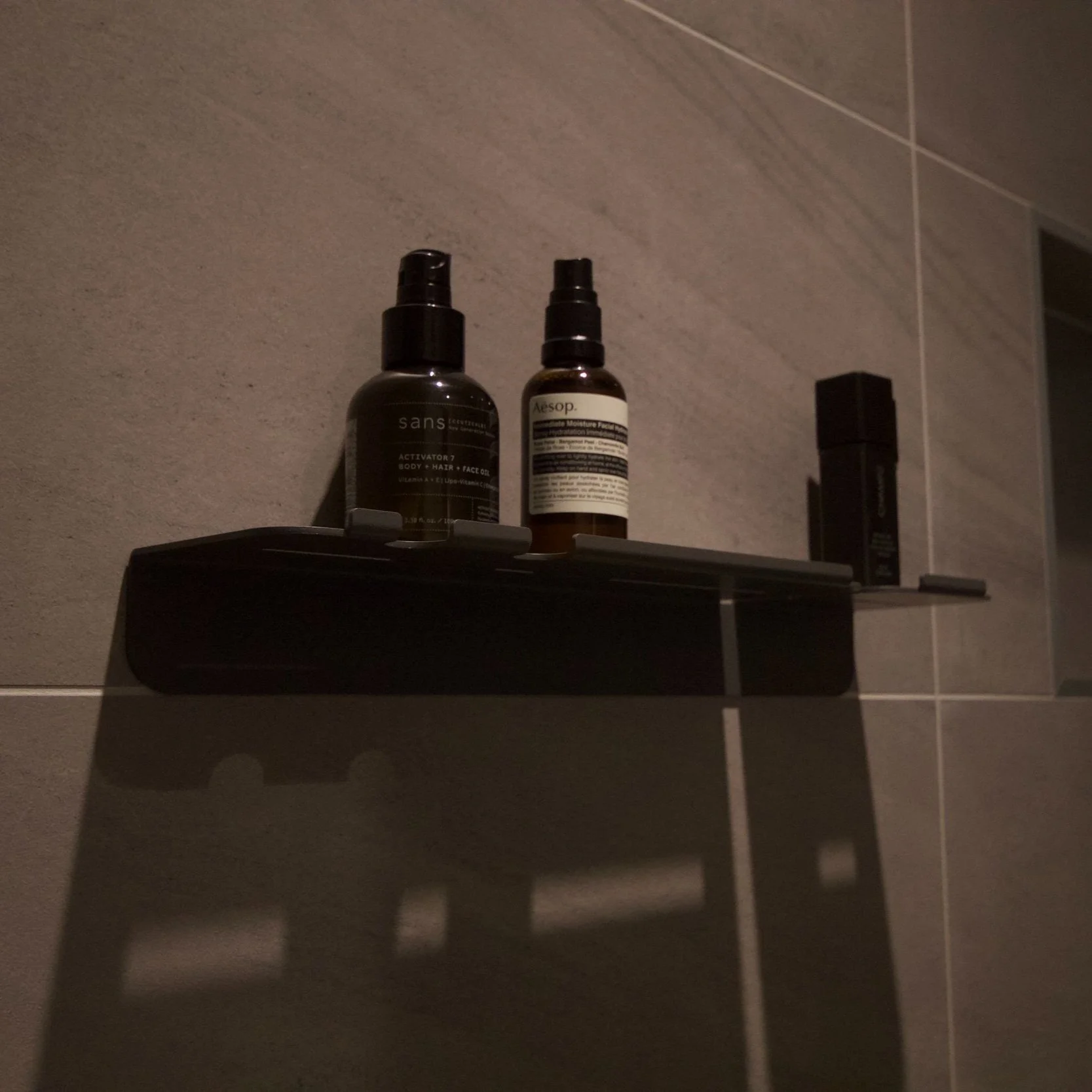 Minimalist grey shower shelf mounted on dark tiles using VHB double-sided tape, featuring a small slot for a toothbrush and two integrated razor holders that double as hooks.