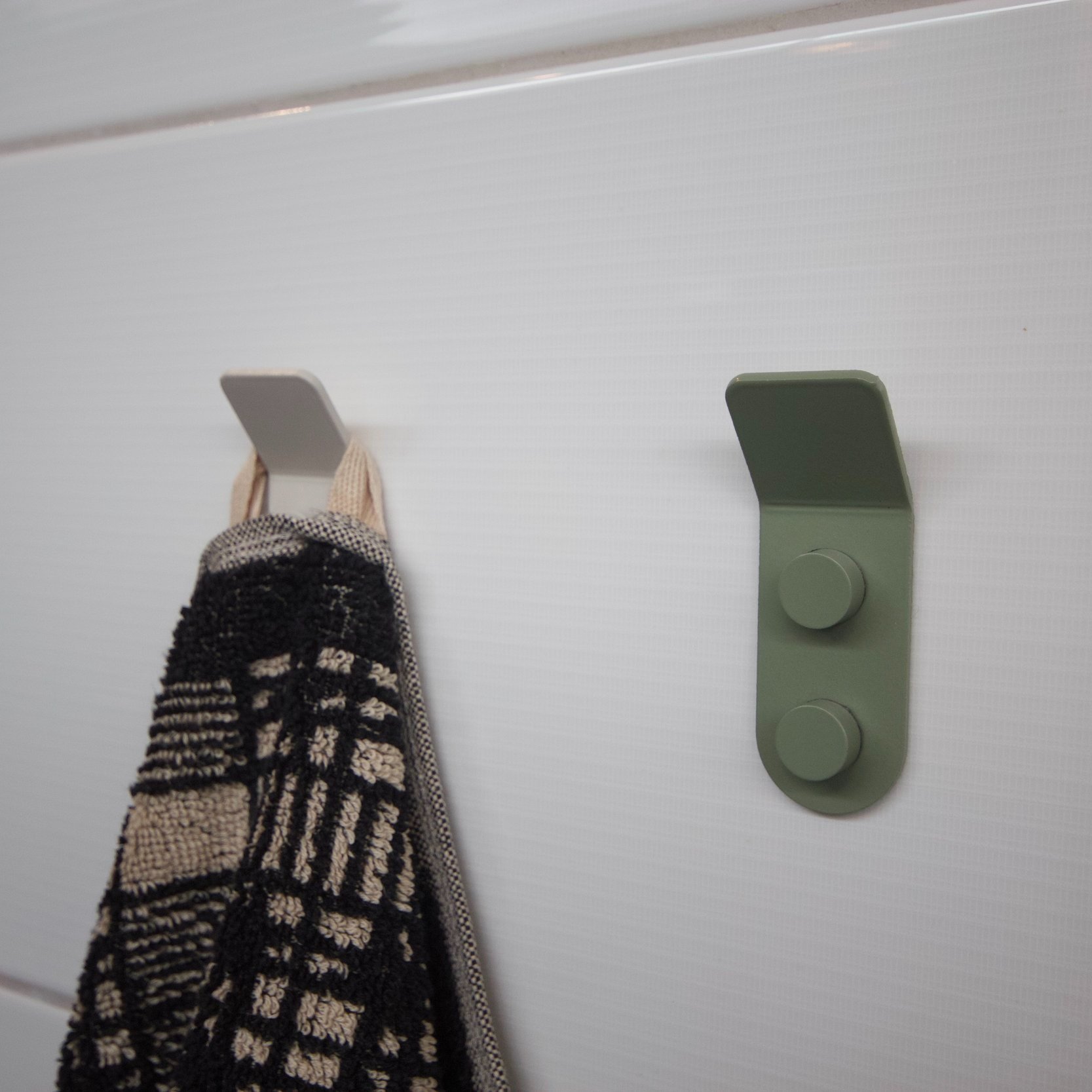 Two minimalist wall hooks mounted on a white tiled wall, one empty gray hook and one green hook holding a black and cream patterned towel.