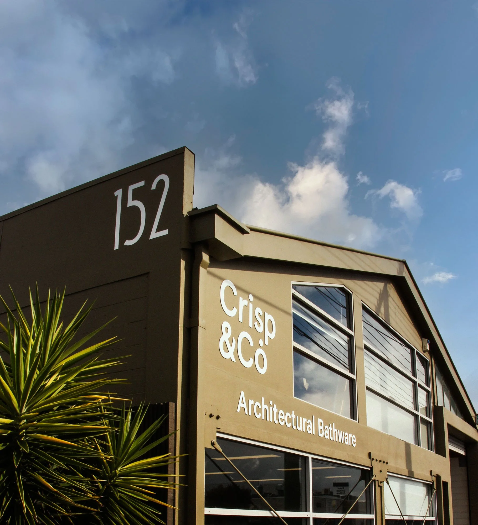 Close-up of a building with large windows and a beige exterior, showing signage for architecture and bathroom products, partially obscured by a plant with spiky green leaves, under a partly cloudy sky.