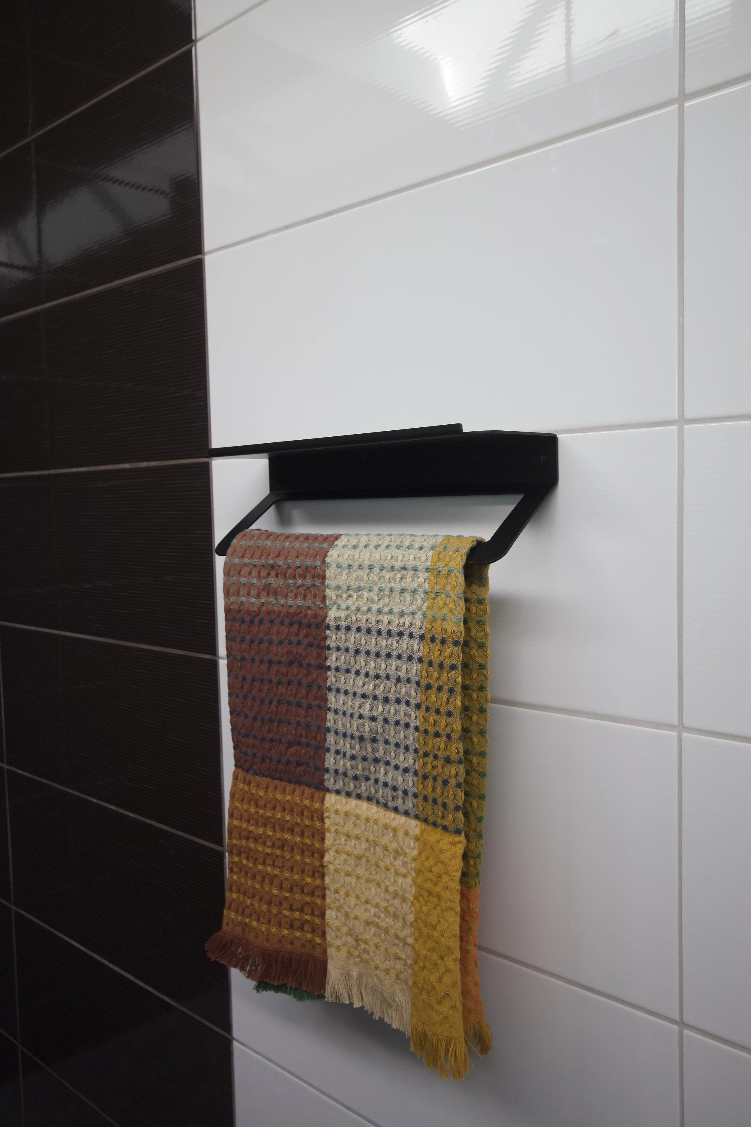 Colorful woven towel hanging on a black wall-mounted towel holder in a bathroom with white and dark brown tiled walls.