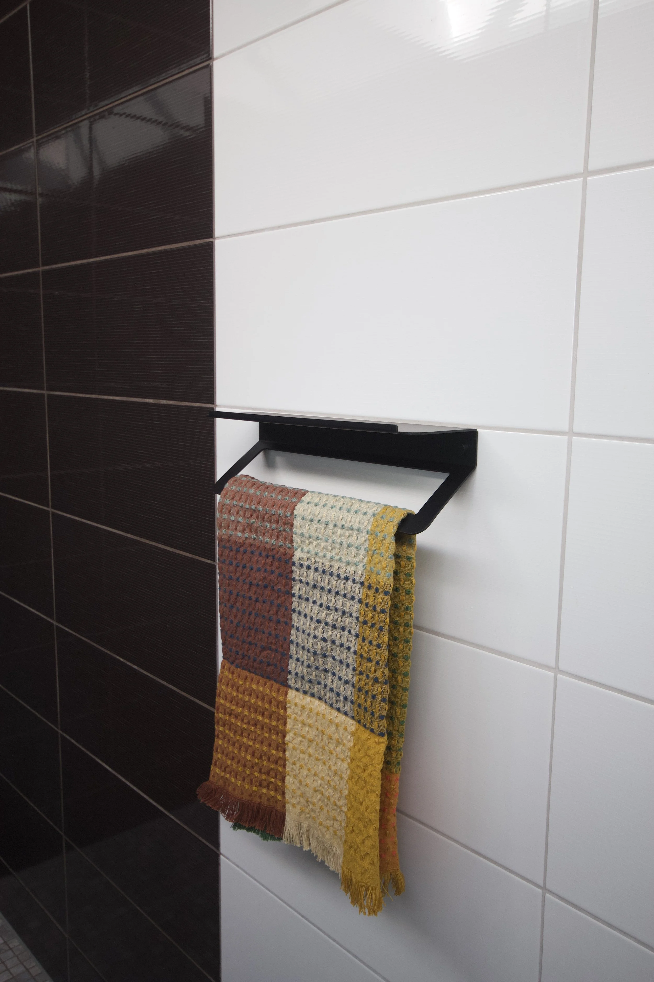 A towel hanging on a black towel rack mounted on a white tiled bathroom wall, with dark brown tiles visible on an adjacent wall.