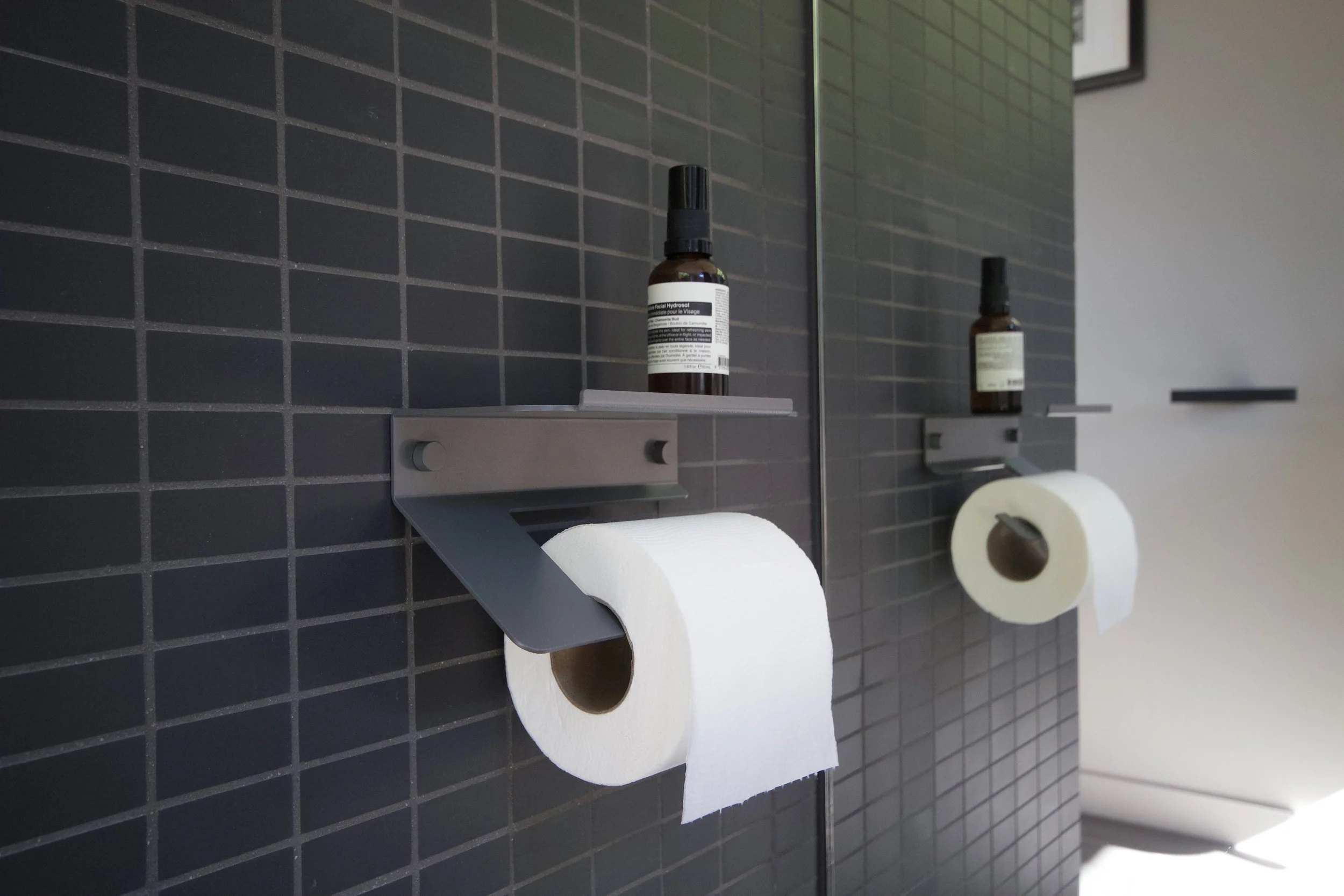 Two bathroom toilet paper holders mounted on a black tiled wall, each holding a roll of toilet paper, with a bottle of spray and a mirror in the background.