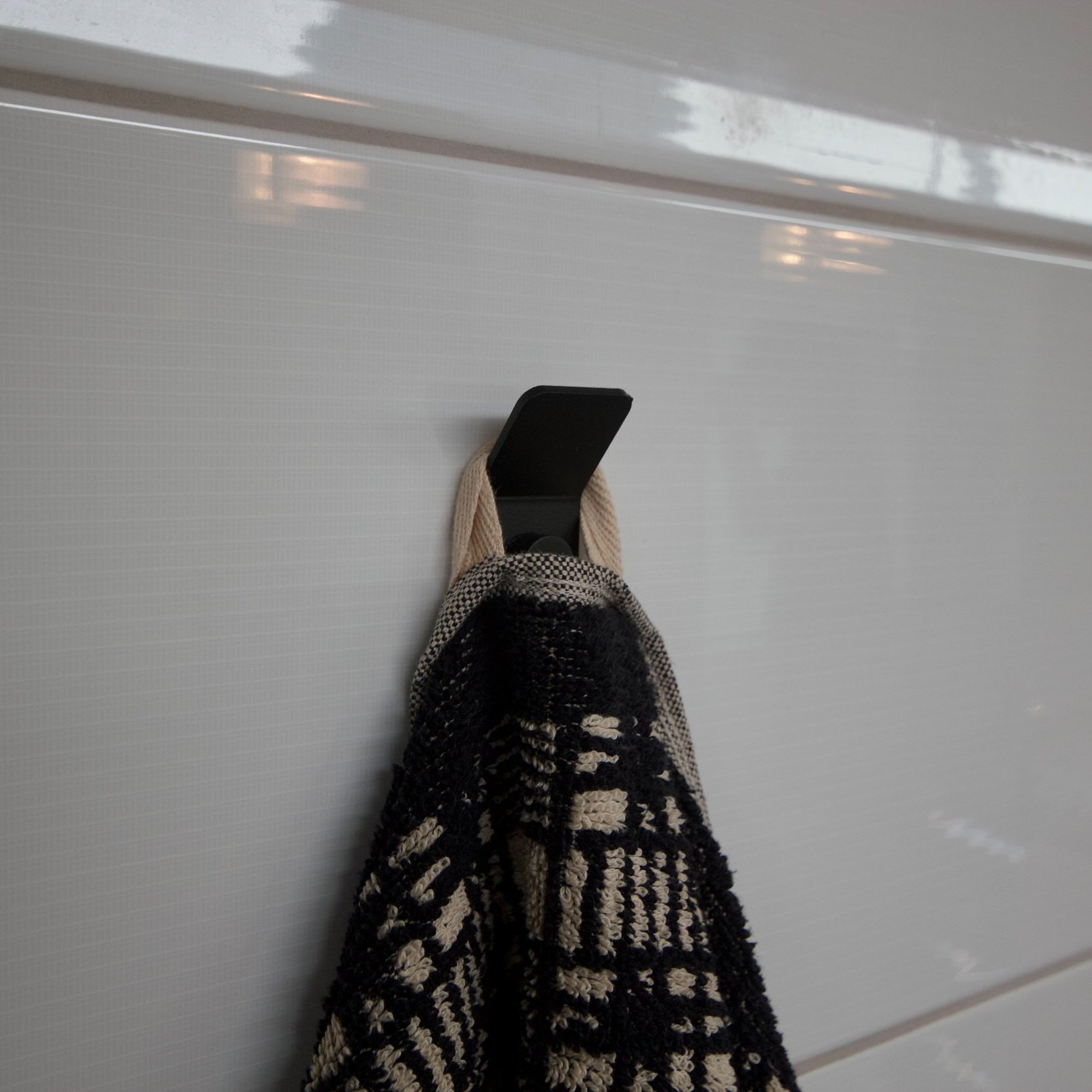 Black aluminium wall hook mounted on a white tiled wall, holding a black and cream patterned towel.