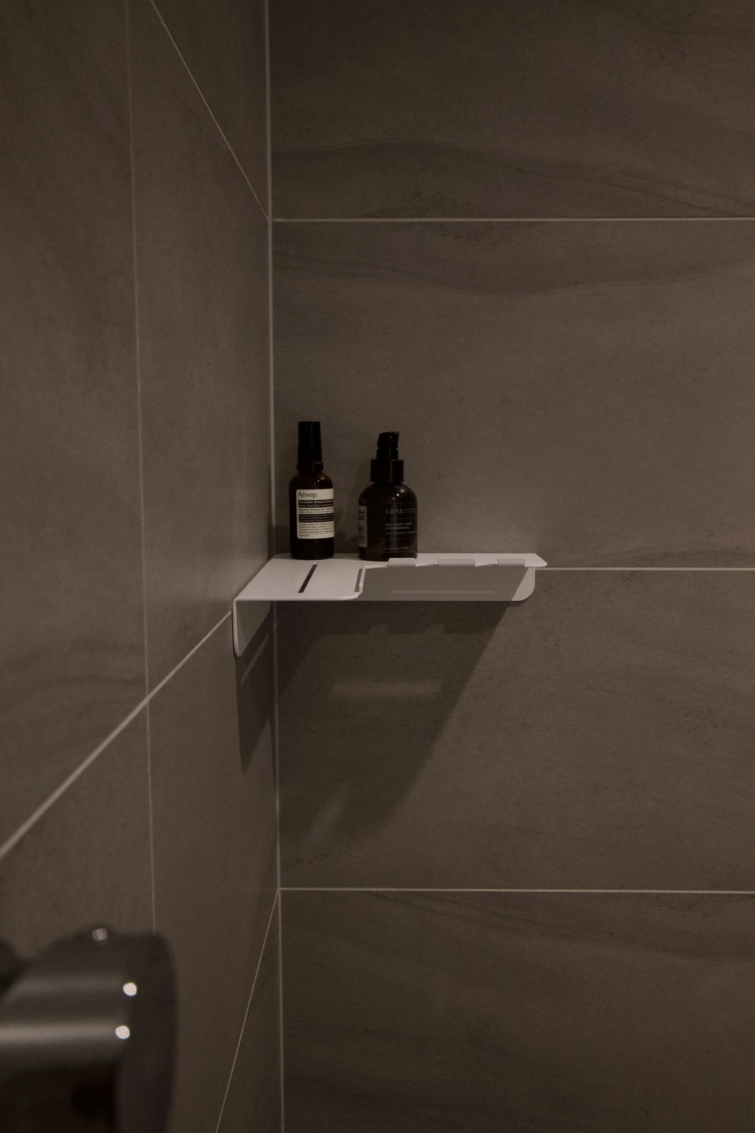 A small white bathroom shelf holding two bottles, one labeled Aesop and the other Sans, mounted on a gray tiled wall.
