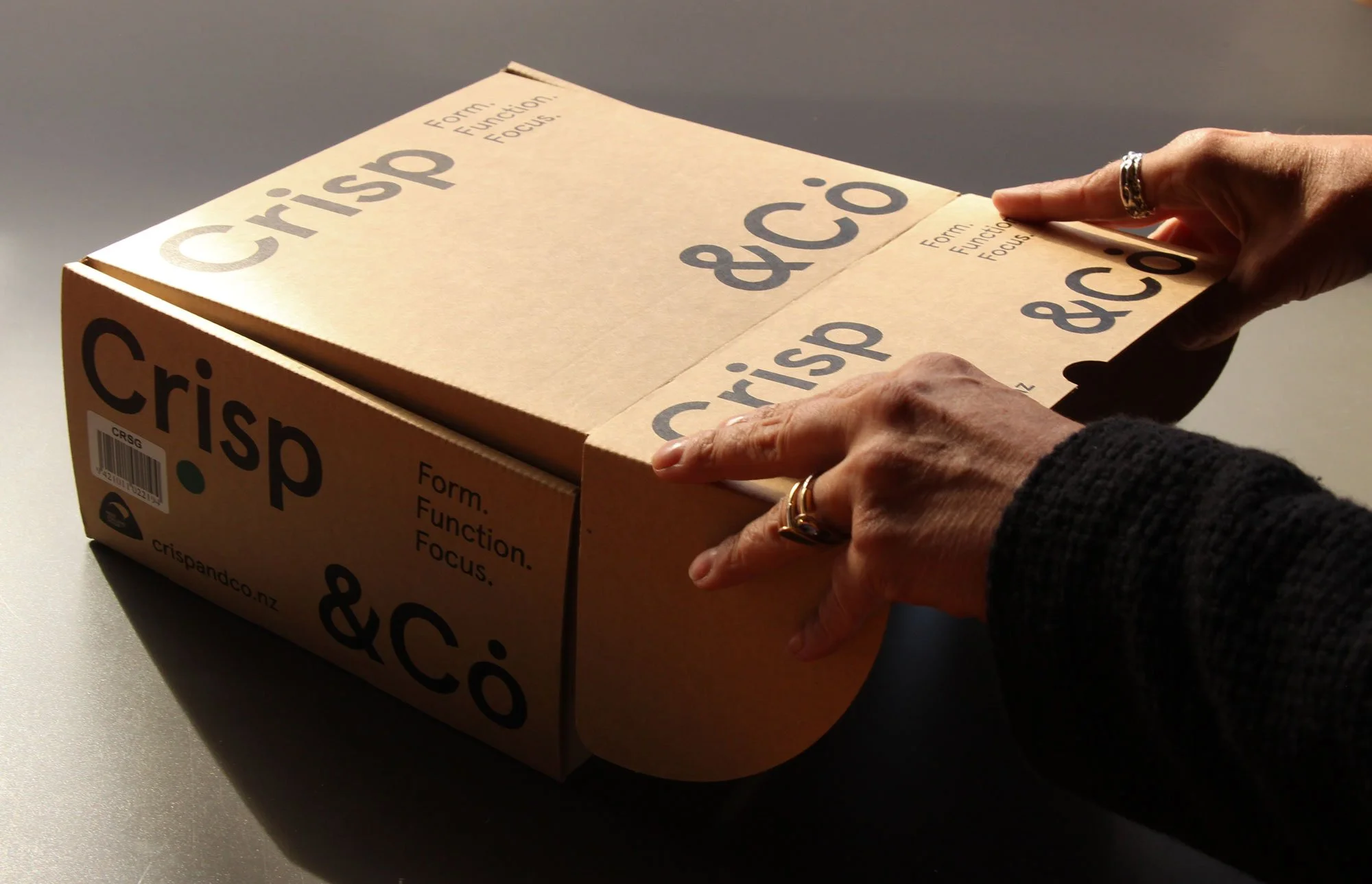 Person opening a cardboard box labeled 'Crisp &Co' with text 'Form. Function. Focus.' on a gray surface, with sunlight casting shadows.