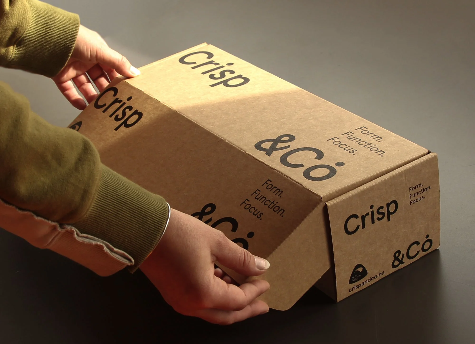 Person holding a cardboard box labeled "Crisp &Co" with their left hand, the person wearing a brown hoodie and light pink pants, standing on a gray surface.