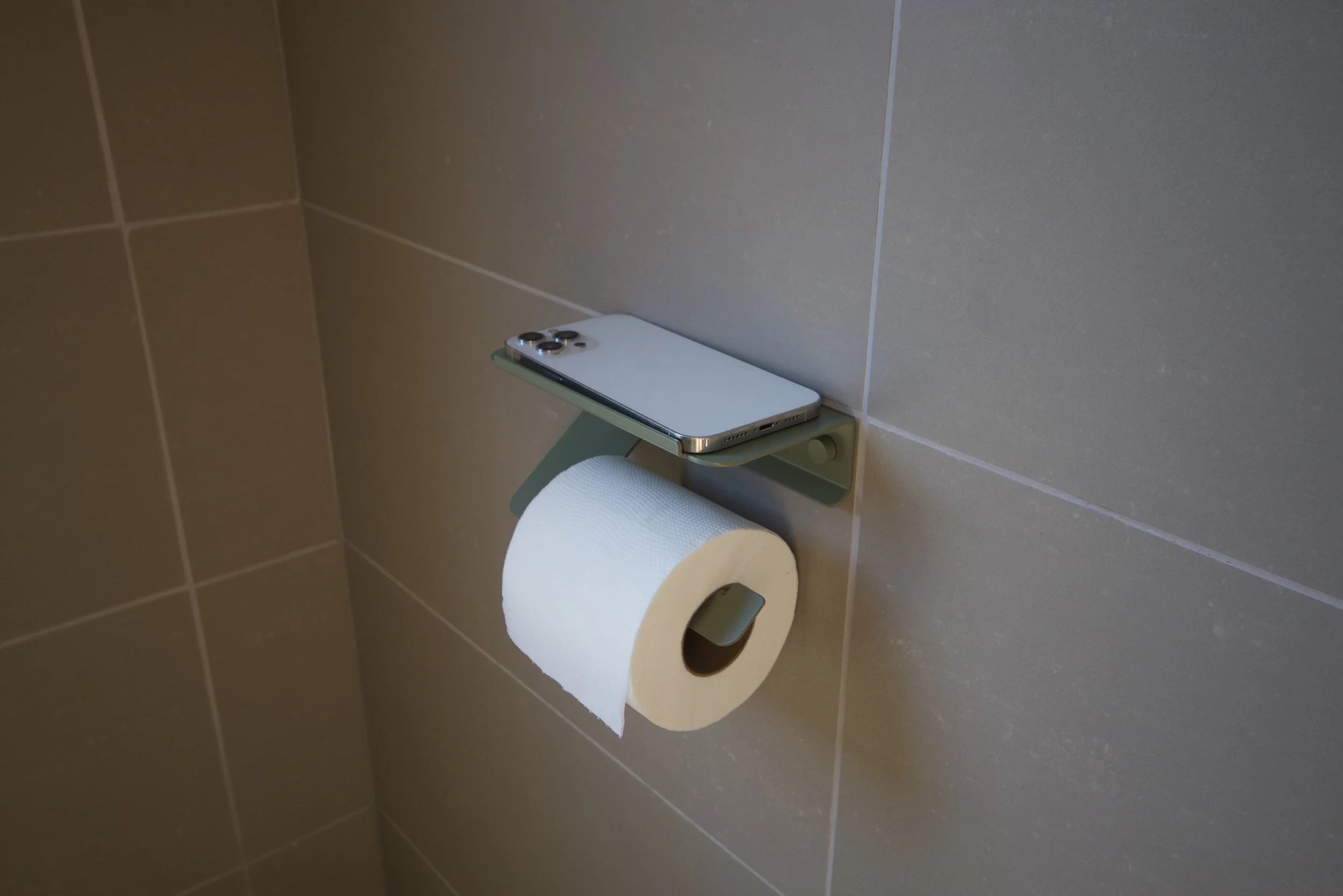A wall-mounted toilet paper holder with a roll of toilet paper and a smartphone resting on top of it, mounted on a tiled wall.