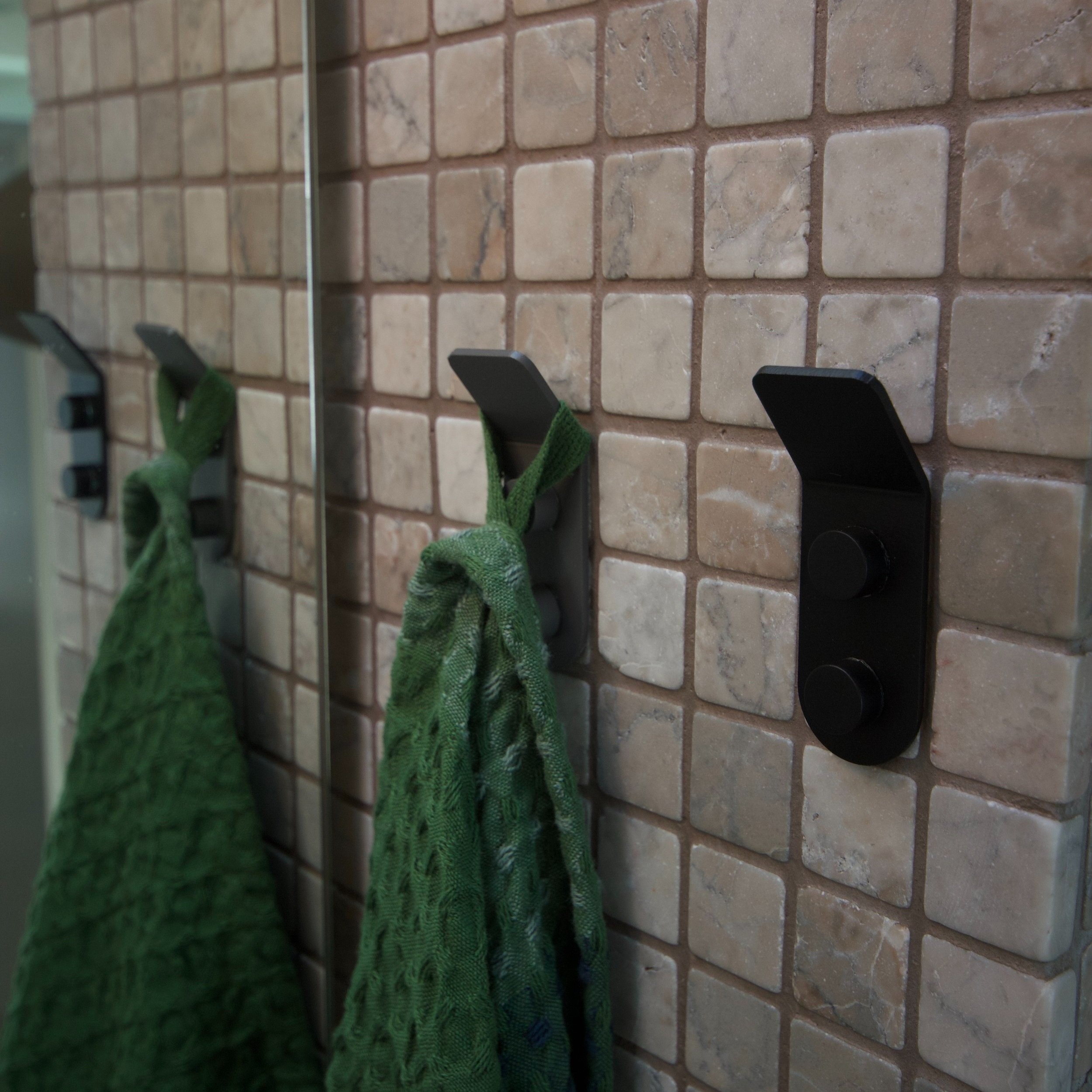 Two minimalist wall hooks mounted on a beige mosaic tiled wall, one holding a green textured towel, with the hooks reflected in a mirror.