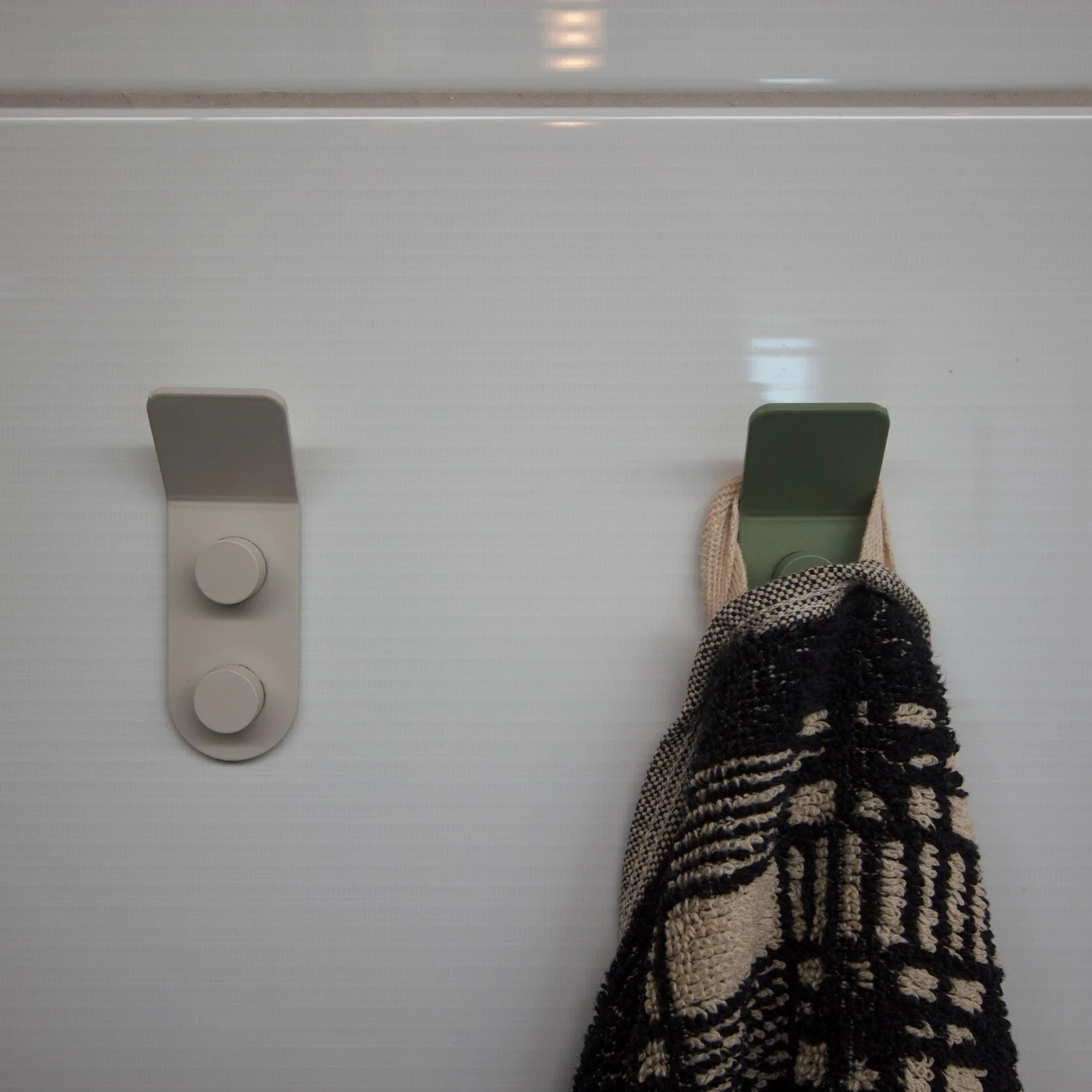 Two minimalist wall hooks mounted on a white tiled wall, one empty gray hook and one green hook holding a black and cream patterned towel.