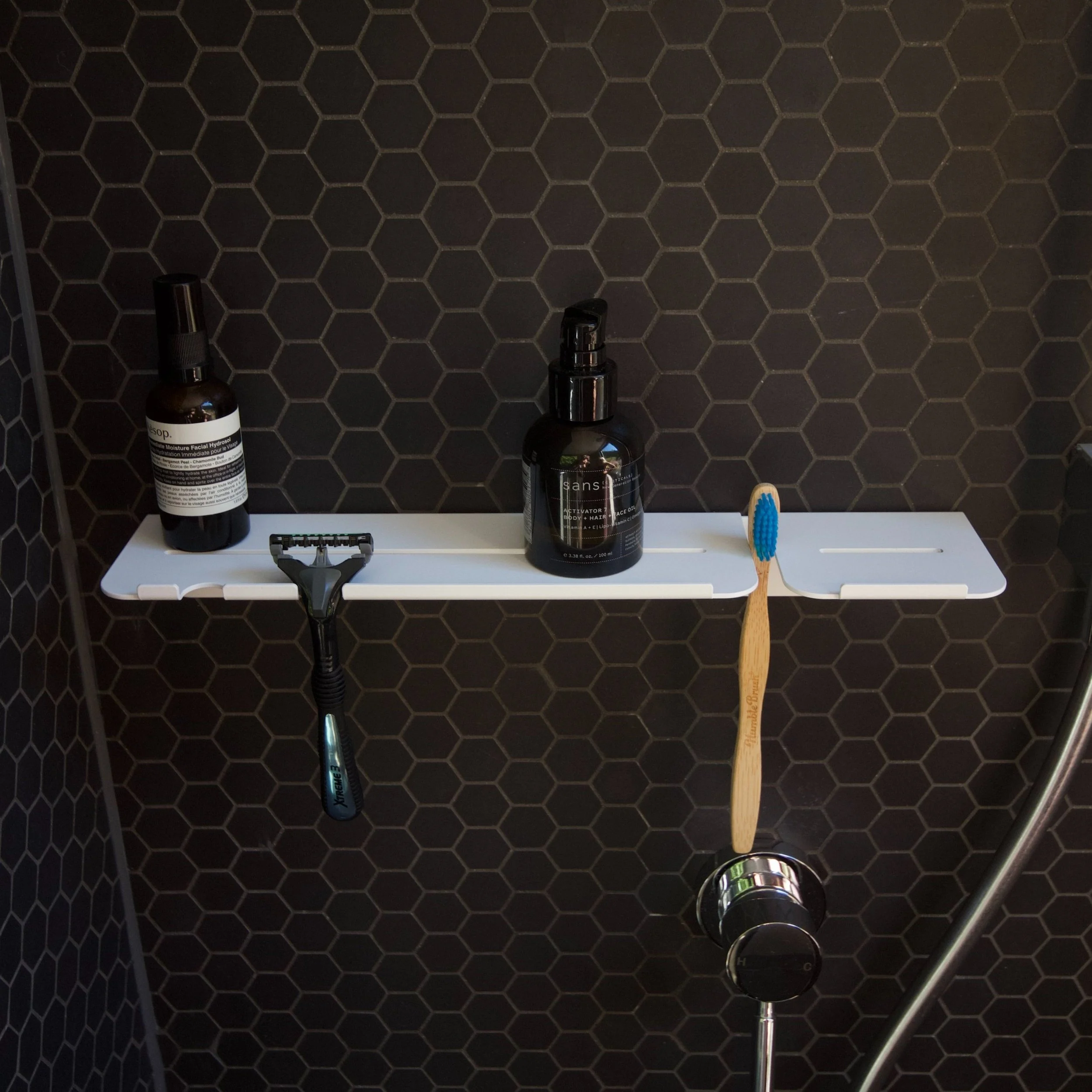 Shelf in a shower with black hexagonal tiles, holding skincare, razor, toothbrush, and shower controls.