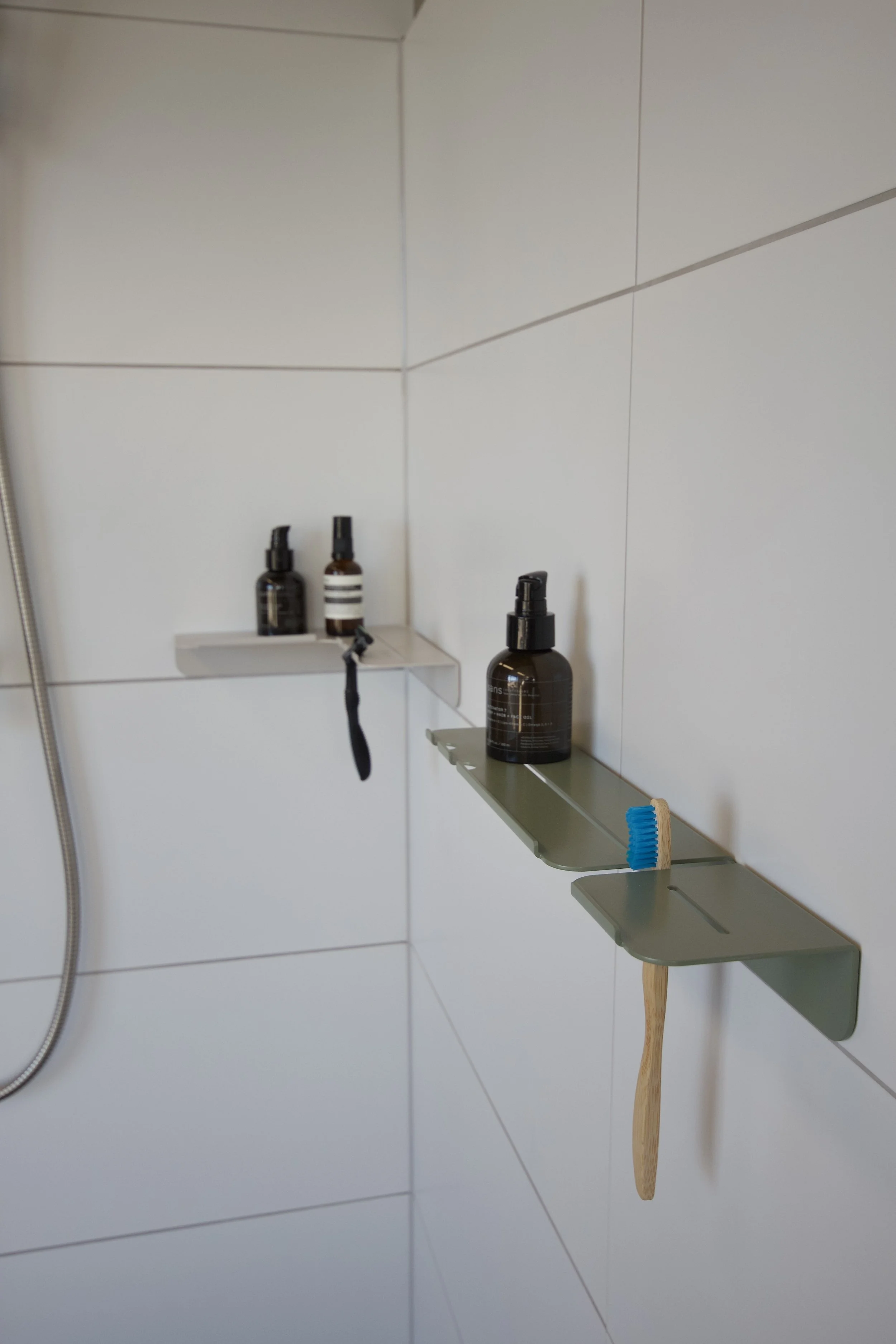 Bathroom shower wall with built-in shelves holding black and brown bottles, a blue scrub brush, and a wooden-handled scraper.