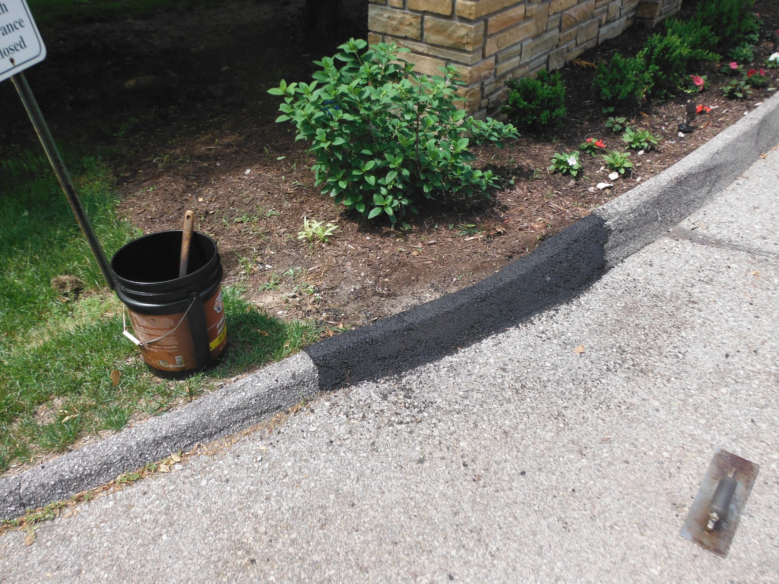 A black bucket with a trowel, black asphalt being laid along the curb, small green plants and shrubs, brick wall, and sidewalk.