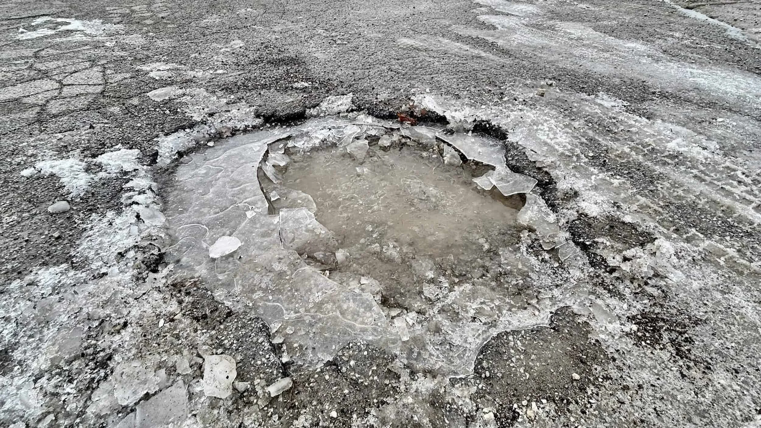 Large winter pothole filled with icy water and broken asphalt on a rough, cracked road surface.