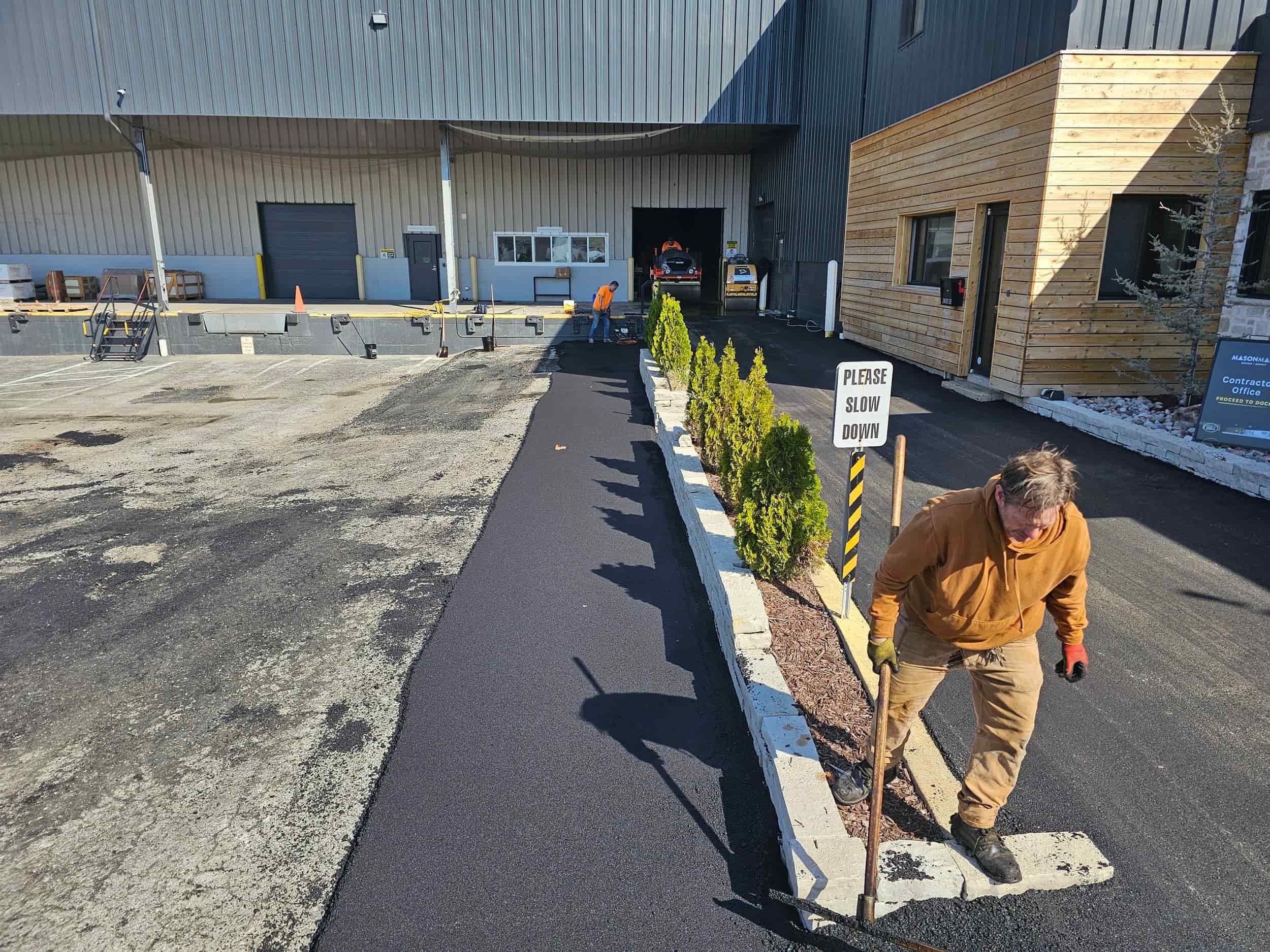 A worker spreads asphalt with a rake near a building, beside a Please slow down sign and some small shrubs.