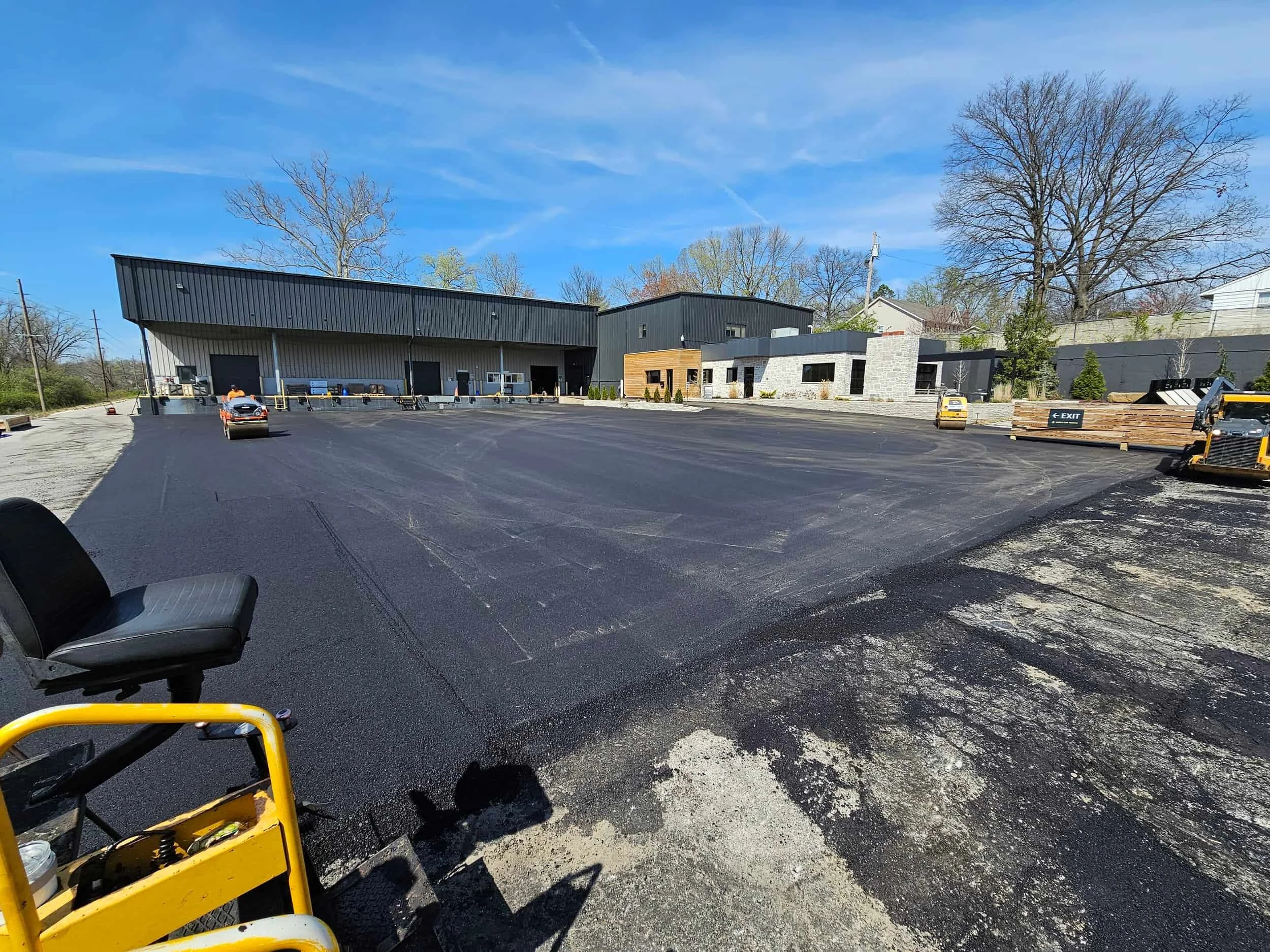 Fresh asphalt being laid in a large parking lot beside a commercial building under a clear blue sky, with paving equipment visible.