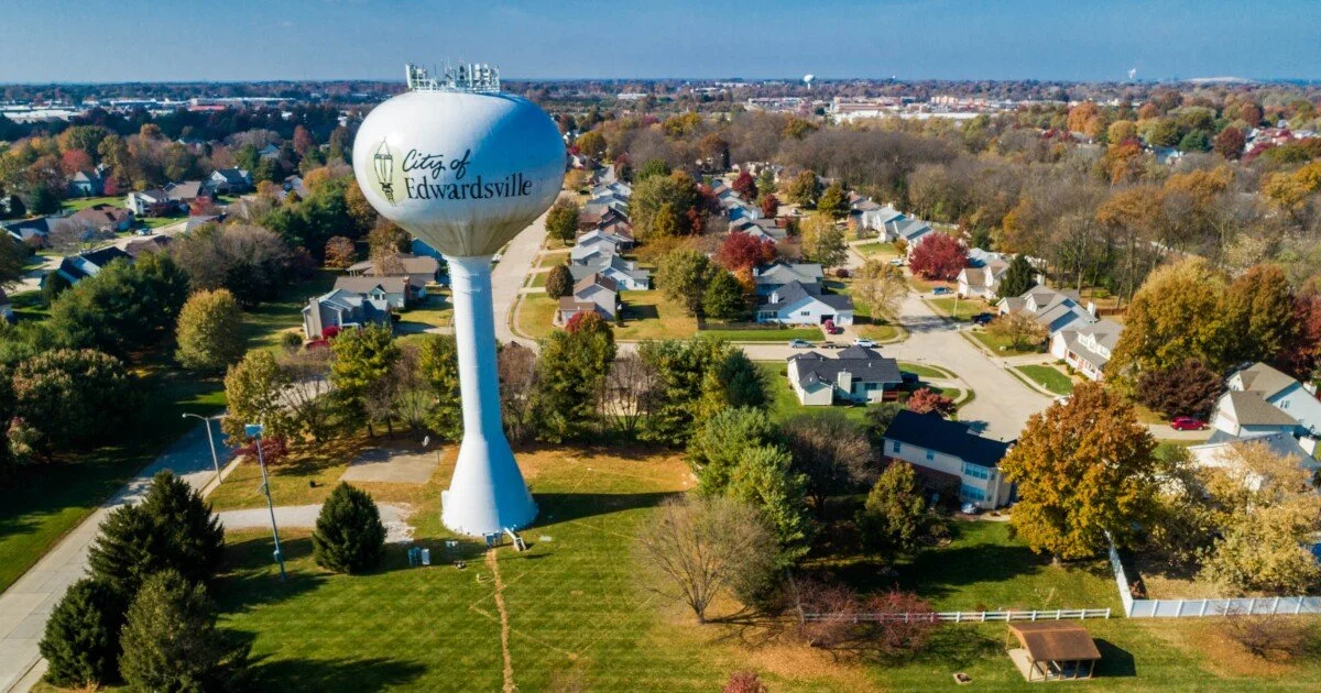A large water tower labeled City of Edwardsville stands amid houses, trees, and green lawns in a suburban neighborhood.