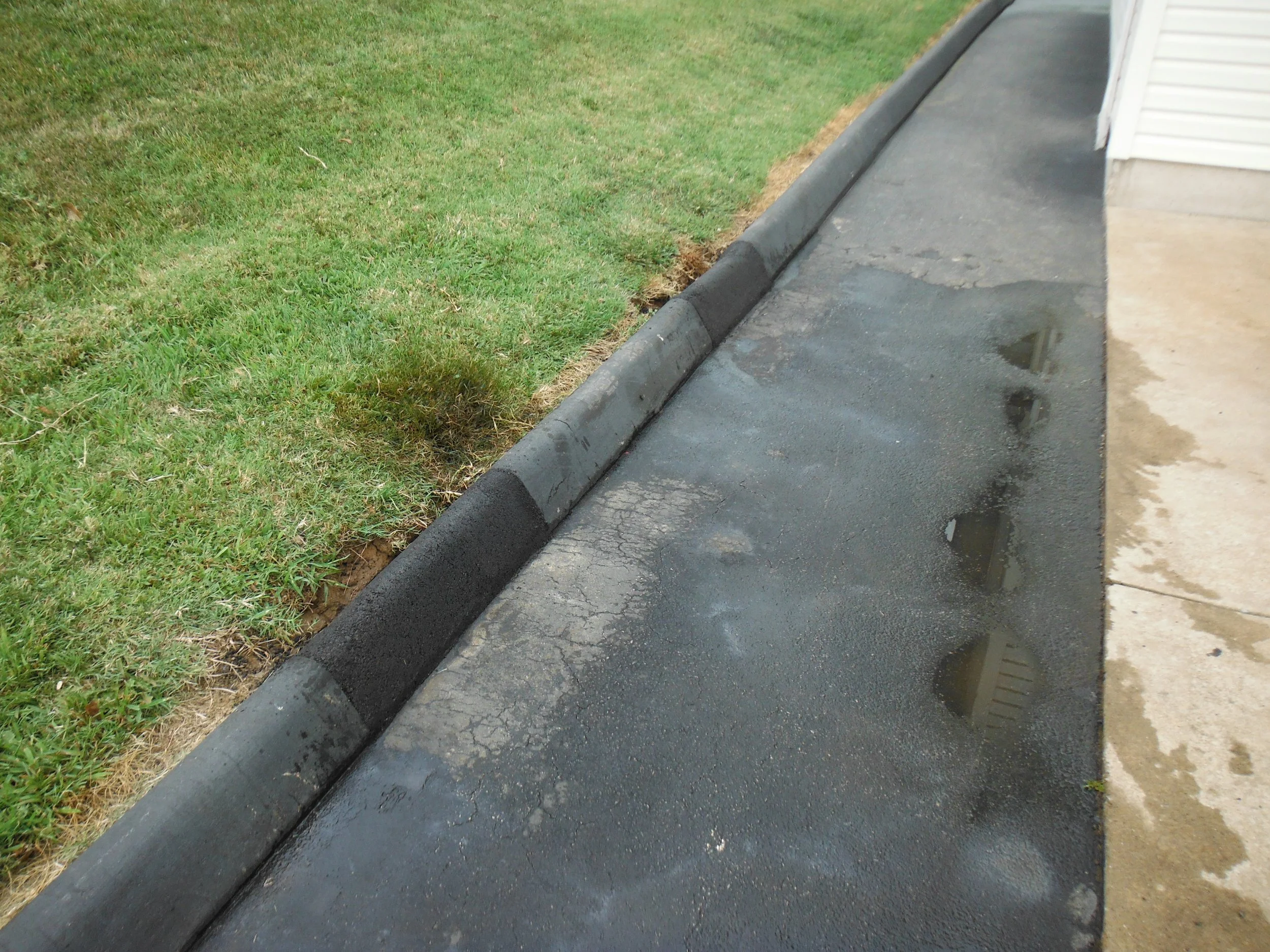 Freshly paved black asphalt sidewalk next to green grass with water puddles and a black concrete curb.