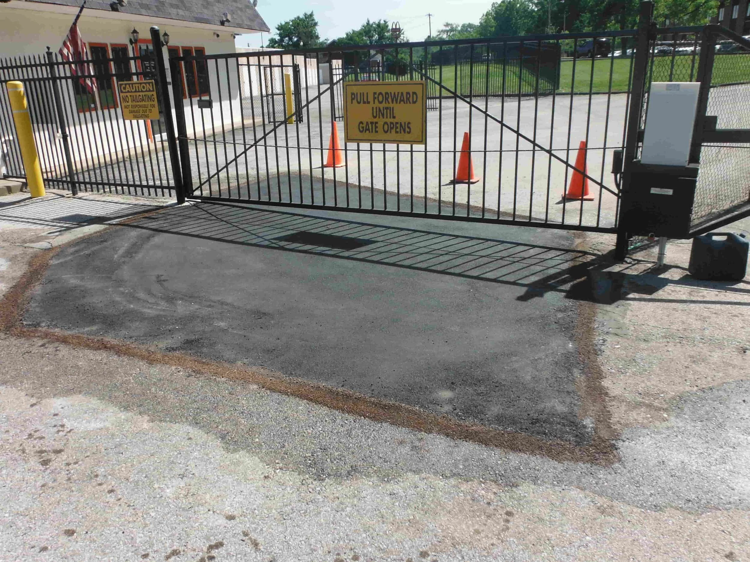 A closed metal gate with warning signs and orange cones; fresh asphalt is patched in front of the gate entrance.