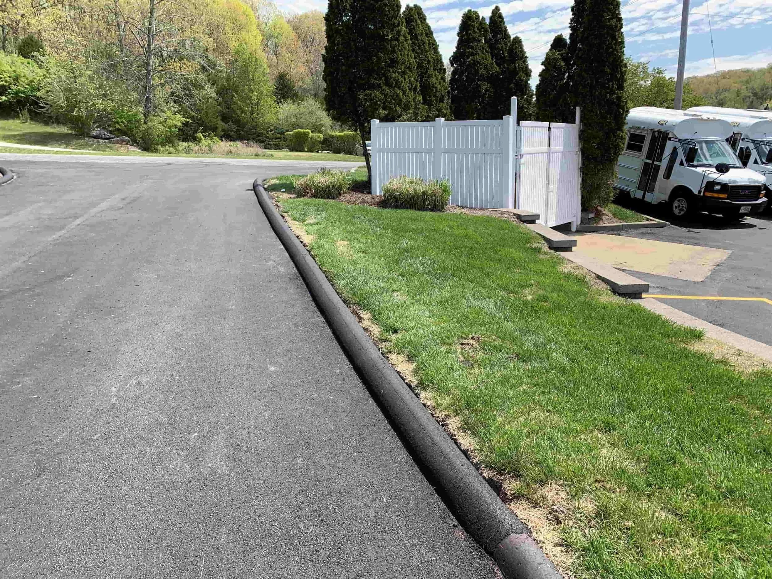 Freshly paved road beside a grassy area, white fence, tall shrubs, and a parked white bus under a partly cloudy sky.
