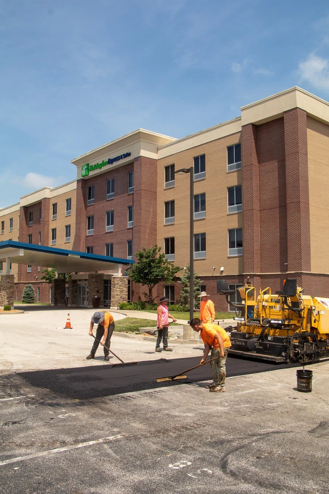 Three workers spread fresh asphalt in a parking lot, with construction vehicles and equipment in the background on a sunny day.