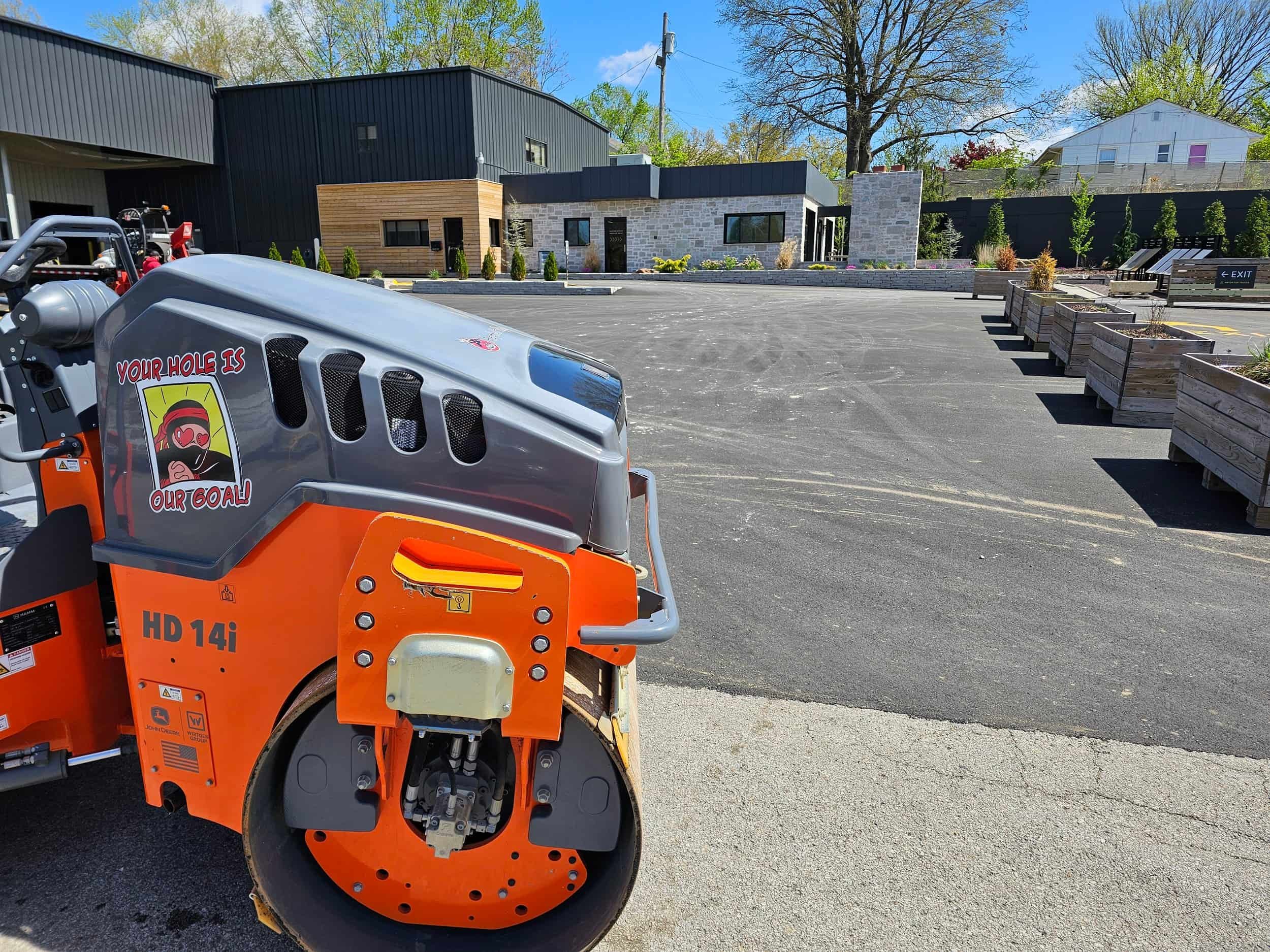 An orange road roller parked on fresh asphalt near a modern building with wooden raised garden beds and clear blue sky above.
