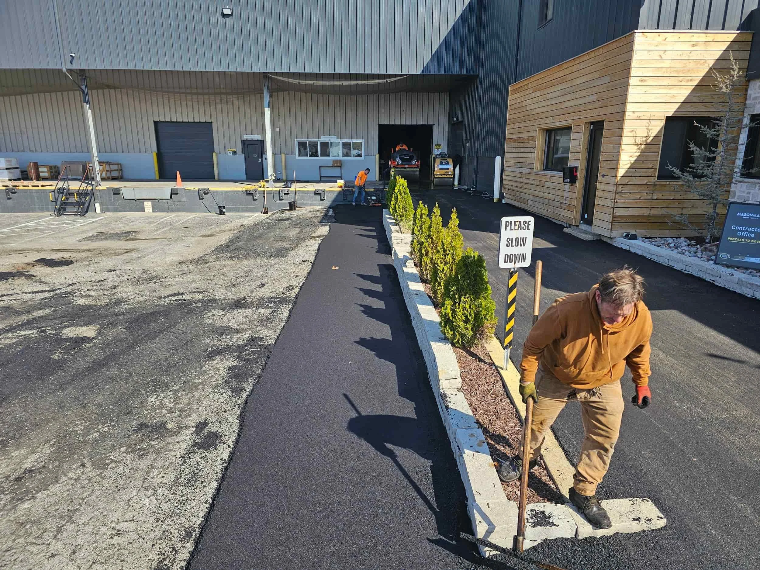 A worker spreads asphalt near a building entrance; a Please Slow Down sign stands beside newly planted shrubs.
