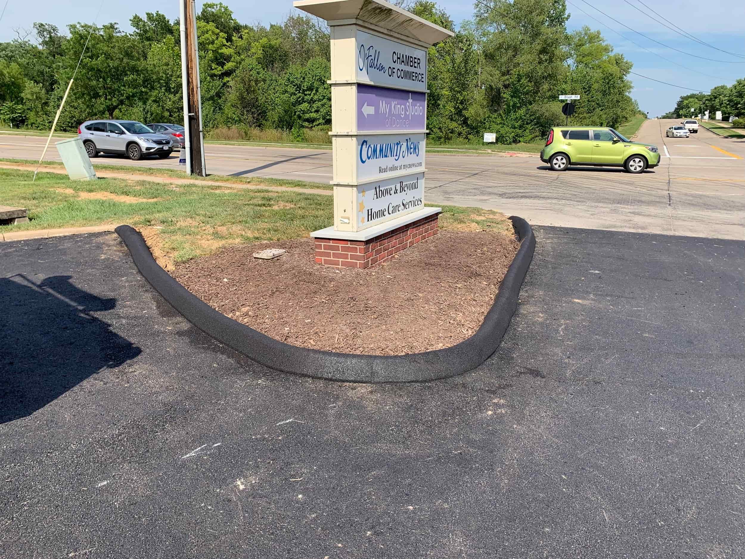 A curved parking lot curb ends abruptly in front of a business sign near a road, with a small green car passing by_ Repaired curb surround business signs at their entrance.