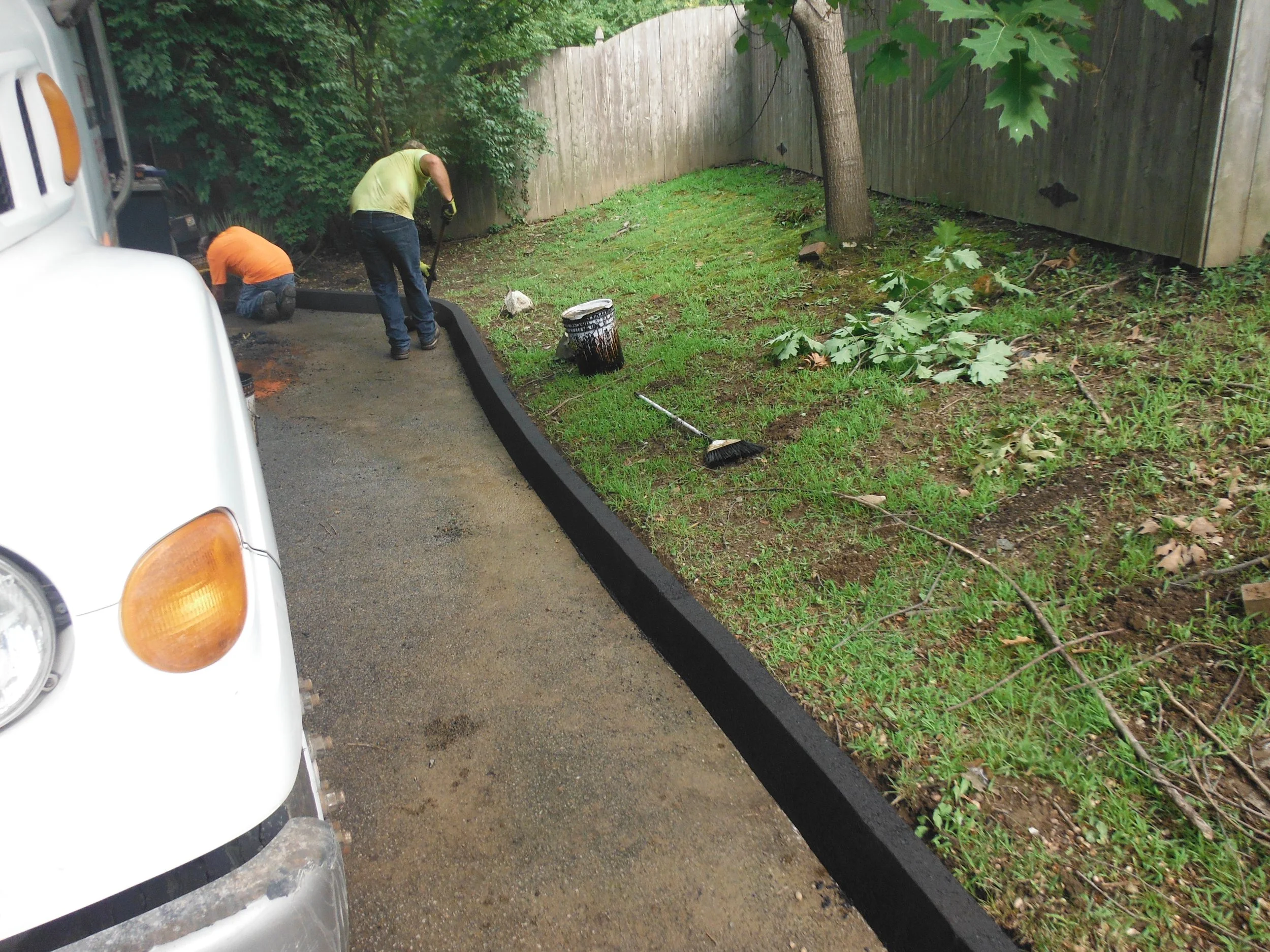 Two workers are installing a new black border along the driveway separating it from the grassy yard. One worker is kneeling while the other is standing with a shovel. Various tools and a paint bucket are nearby. The driveway is partially paved, with 