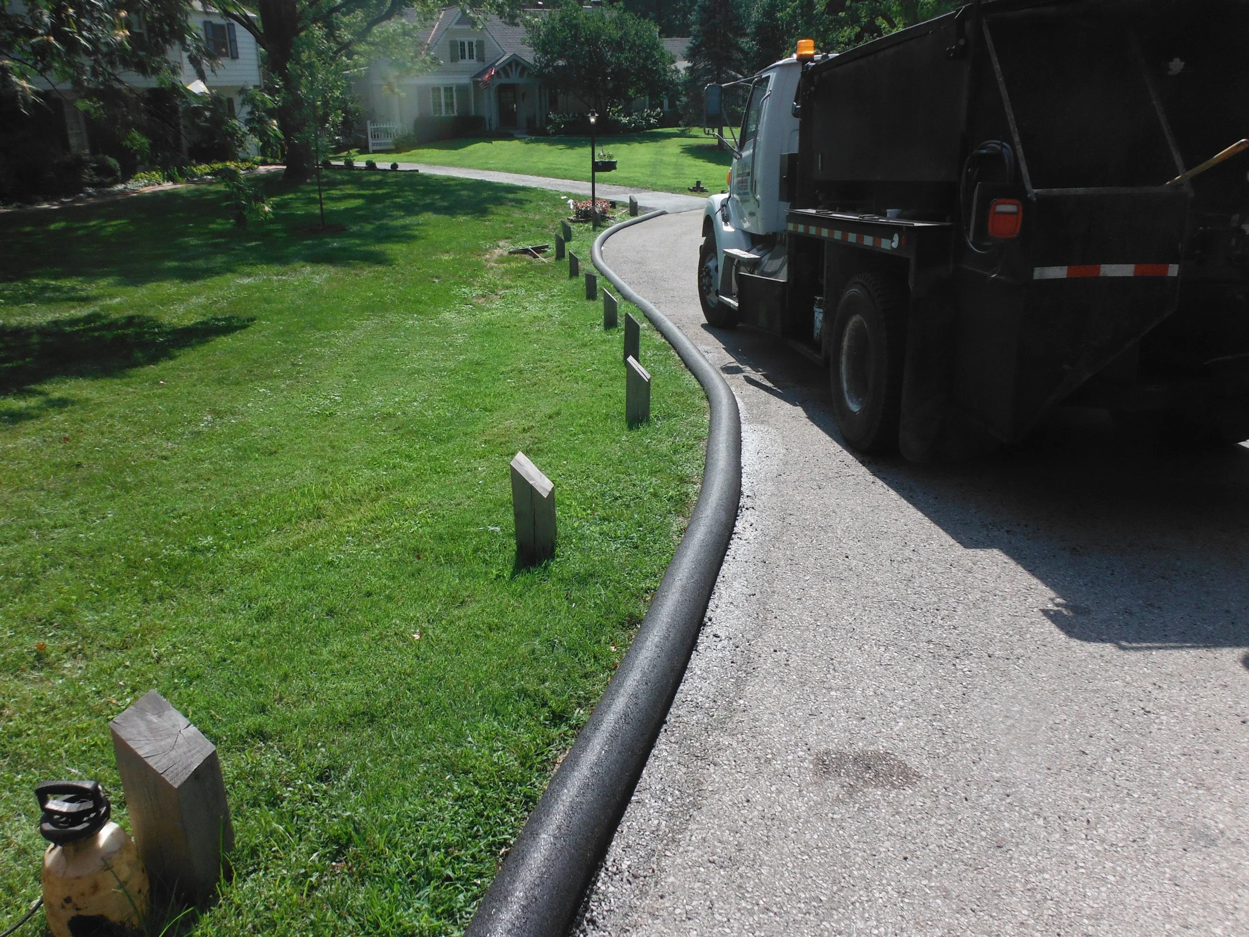 A street with a truck and a line of evenly spaced wooden posts along the sidewalk, with grass and residential houses in the background.